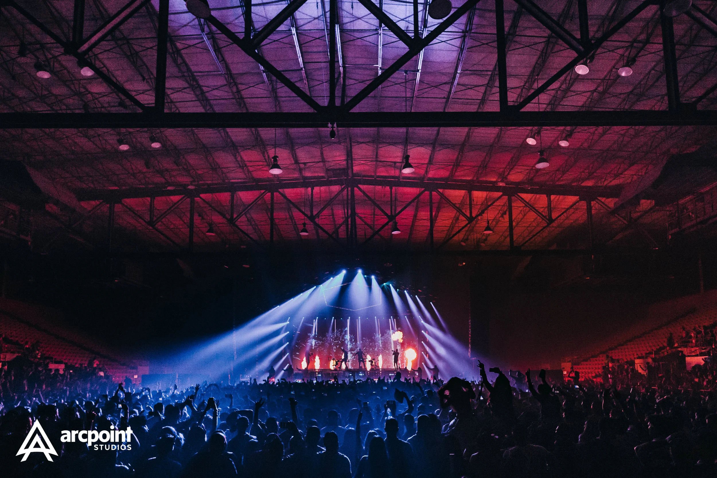 A large indoor concert or music event with a crowd of people facing a brightly lit stage. The stage features performers with colorful lighting effects, including blue and purple beams, with a background of bright screens displaying visuals. The venue