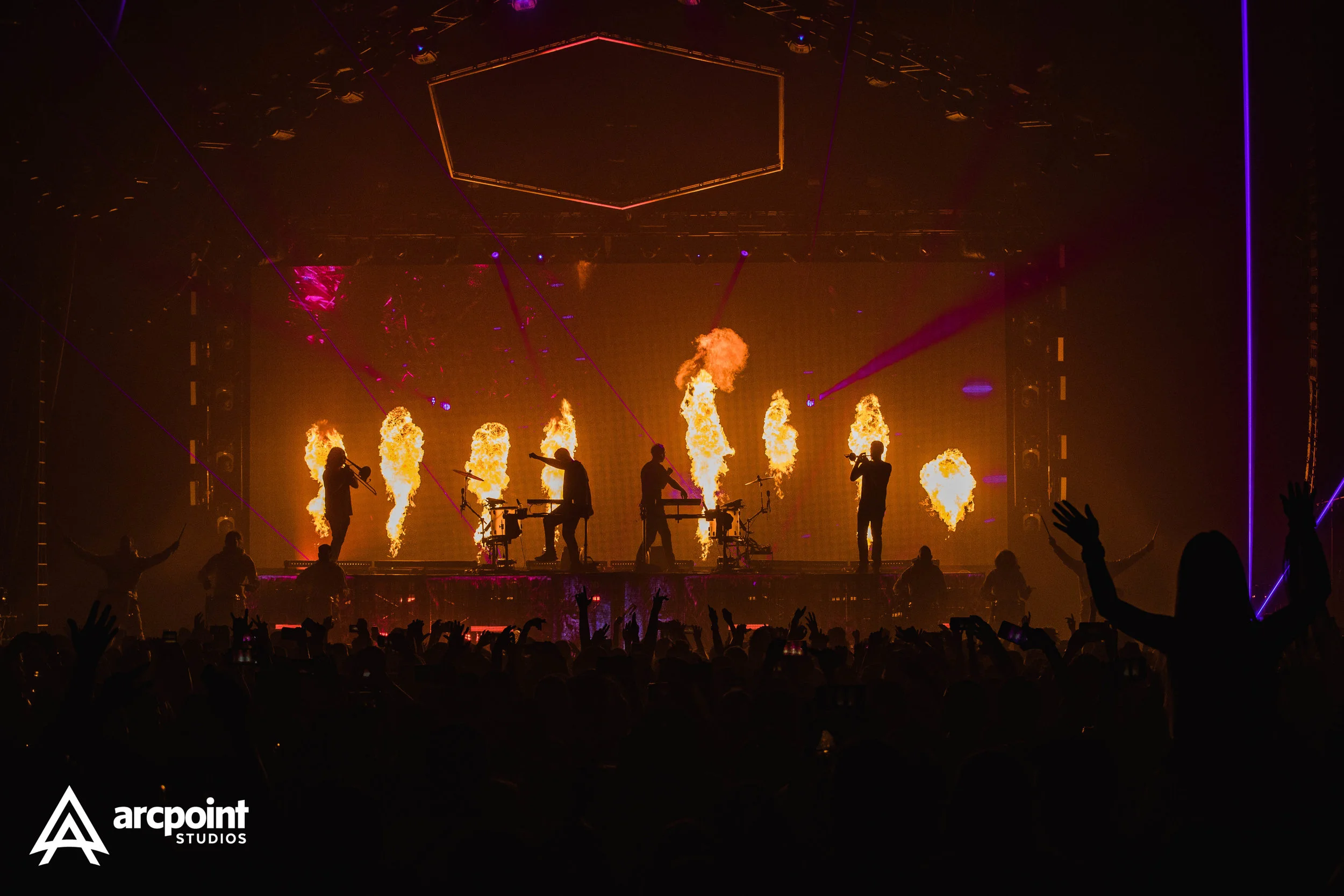 Concert stage with performers and pyrotechnics in the background, audience with raised hands in the foreground, and Arcapoint Studios logo in the bottom left corner.
