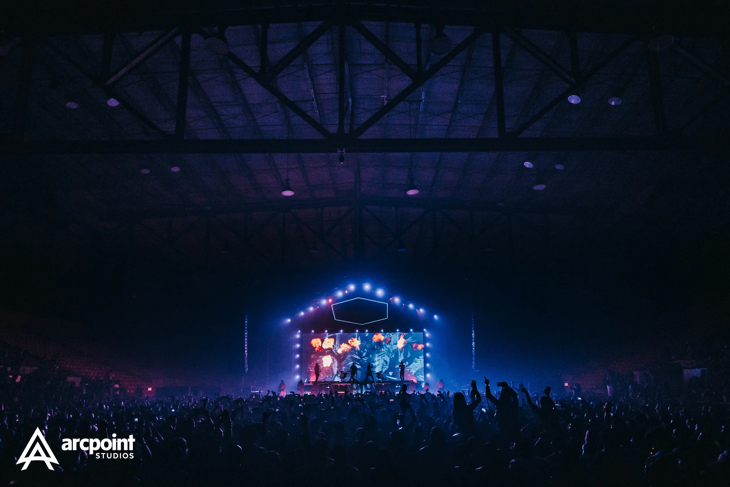 A concert at a large indoor arena with a stage illuminated by colorful lights and a digital screen displaying visual effects, with a crowd of people enjoying the performance.