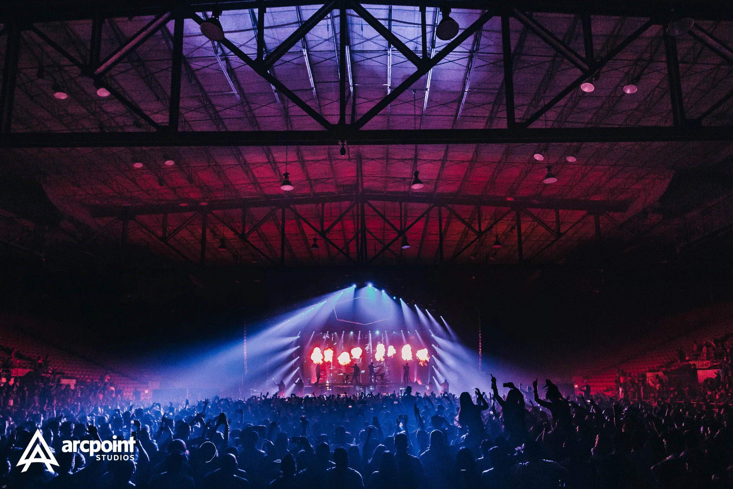 Concert crowd in a dark indoor arena with colorful stage lighting and performers on stage.