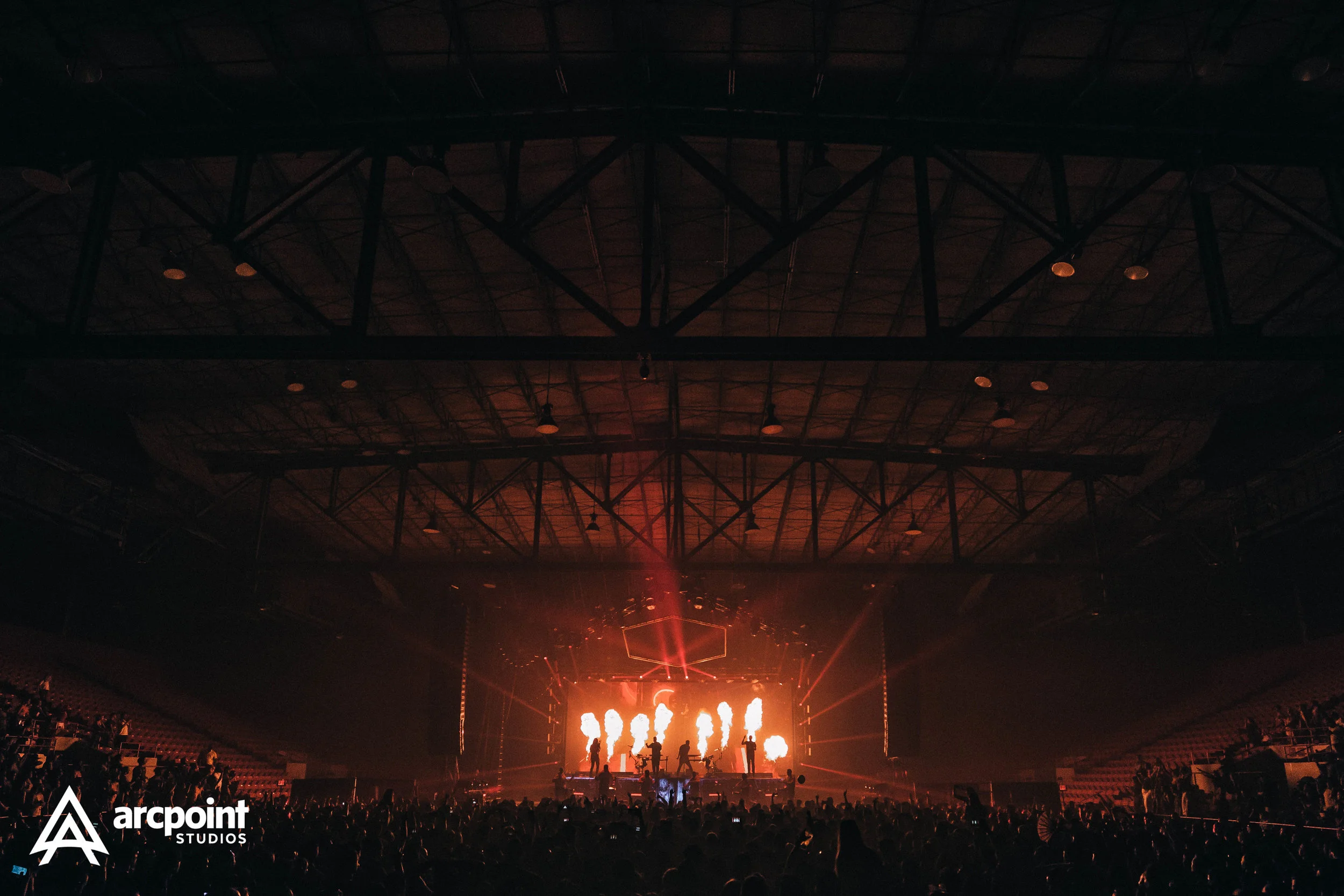 A concert stage illuminated with bright flames and performance lights, with a large audience in front, inside an indoor arena with a high ceiling and structural beams. The logo for 'arcpoint STUDIOS' appears in the bottom left corner.