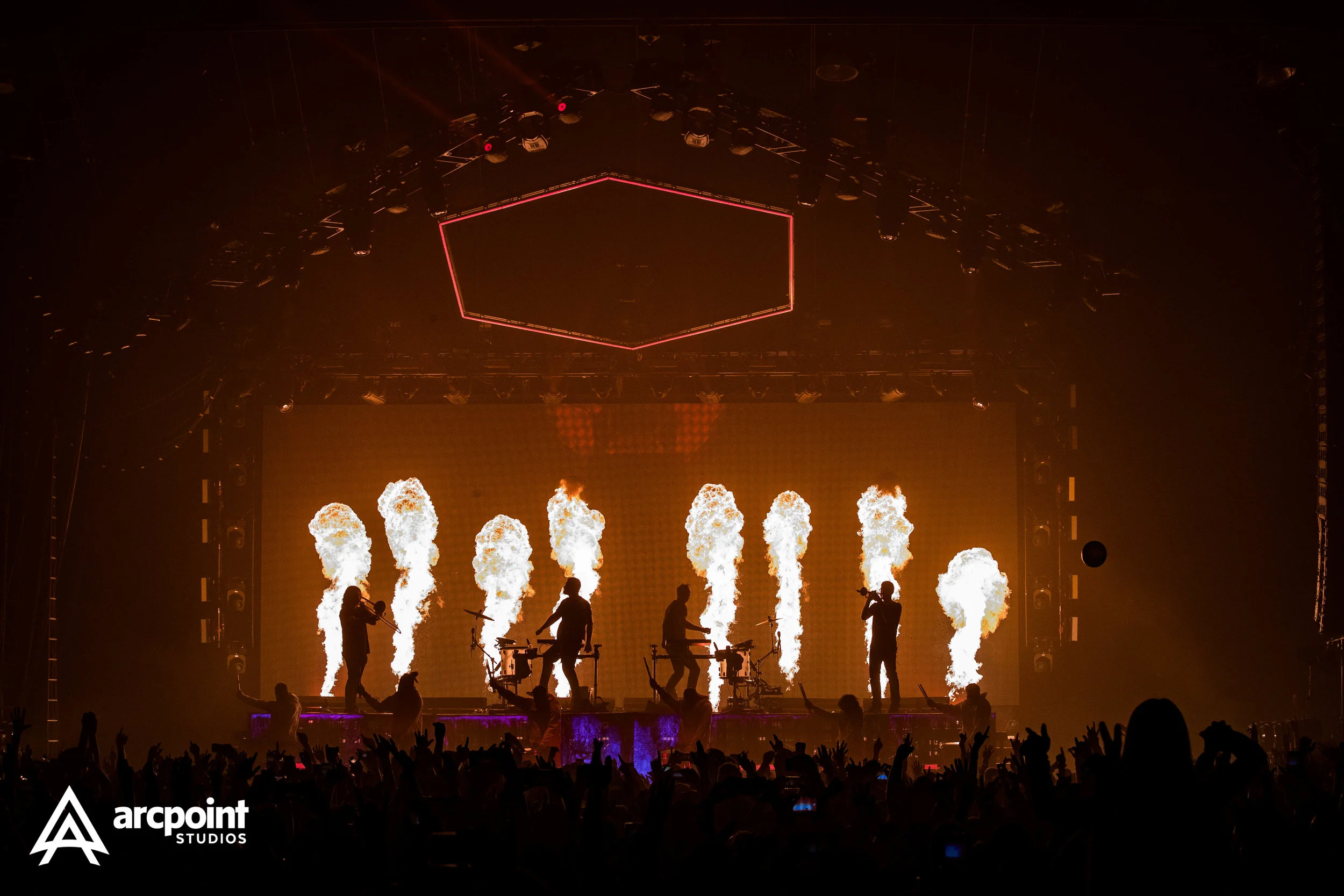 Musicians on stage with pyrotechnics and flames, audience with raised hands, concert lighting, and screen display.