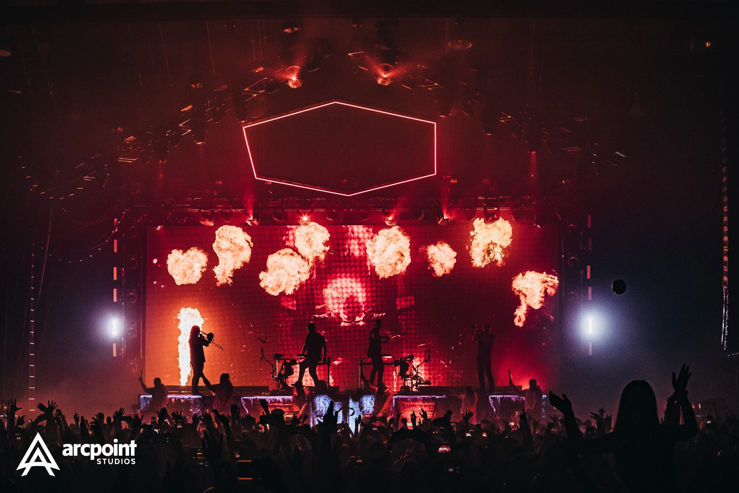 A concert stage with musicians, pyrotechnics, and bright red and white lighting, with an audience in the foreground.