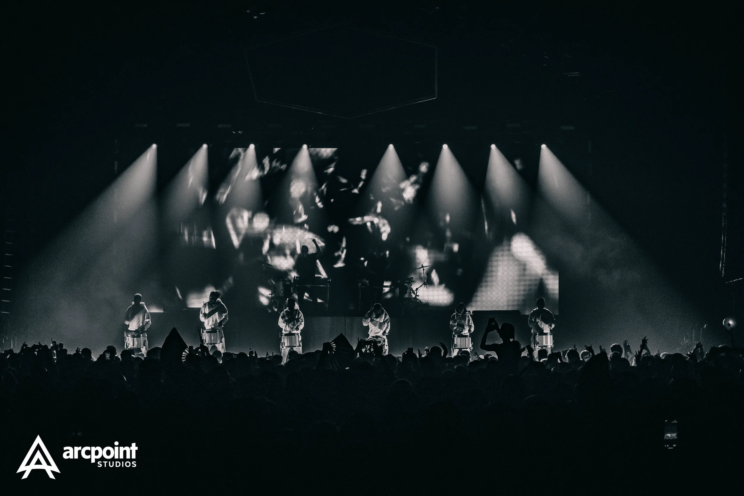 A black and white photo of a concert stage with six performers playing drums, with spotlights shining down on them. A large audience is visible in front of the stage, some holding up their phones.