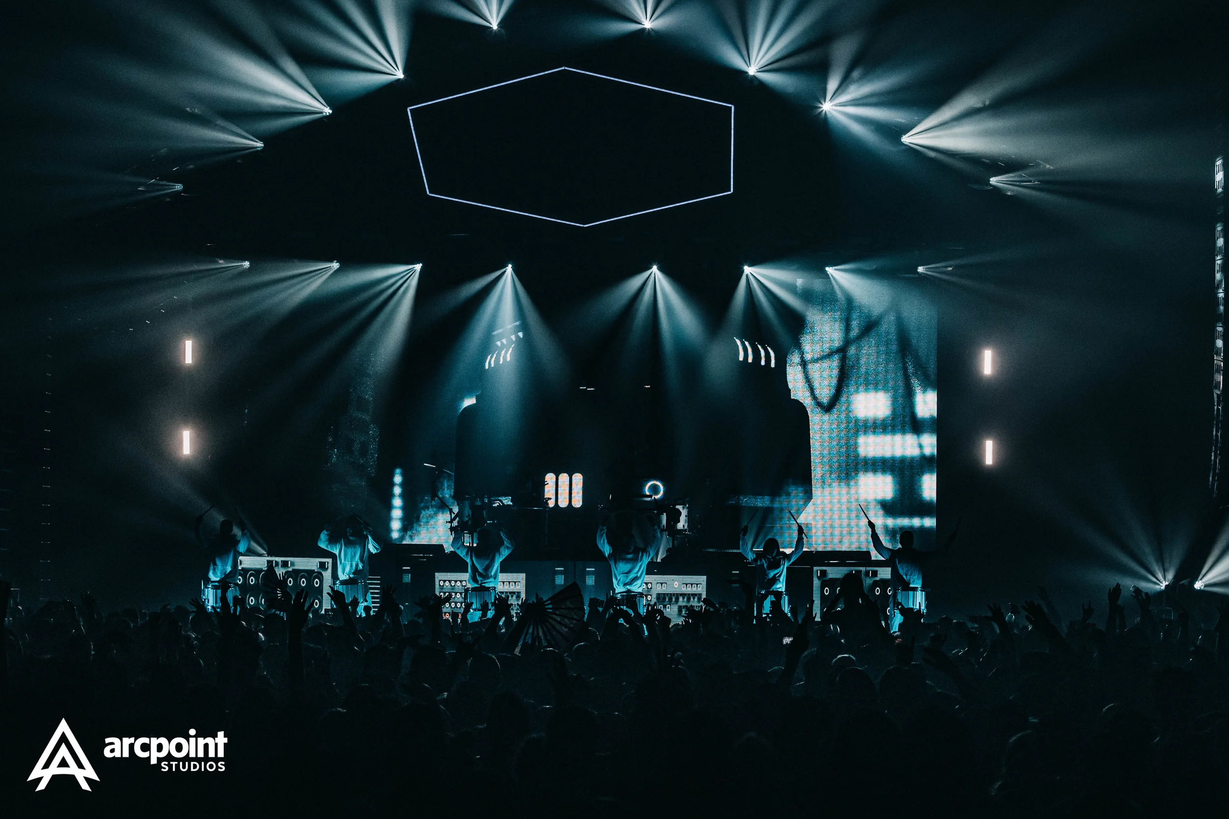 A dark concert scene with silhouettes of performers on stage, illuminated by blue and white lights, with a crowd of fans in the foreground.