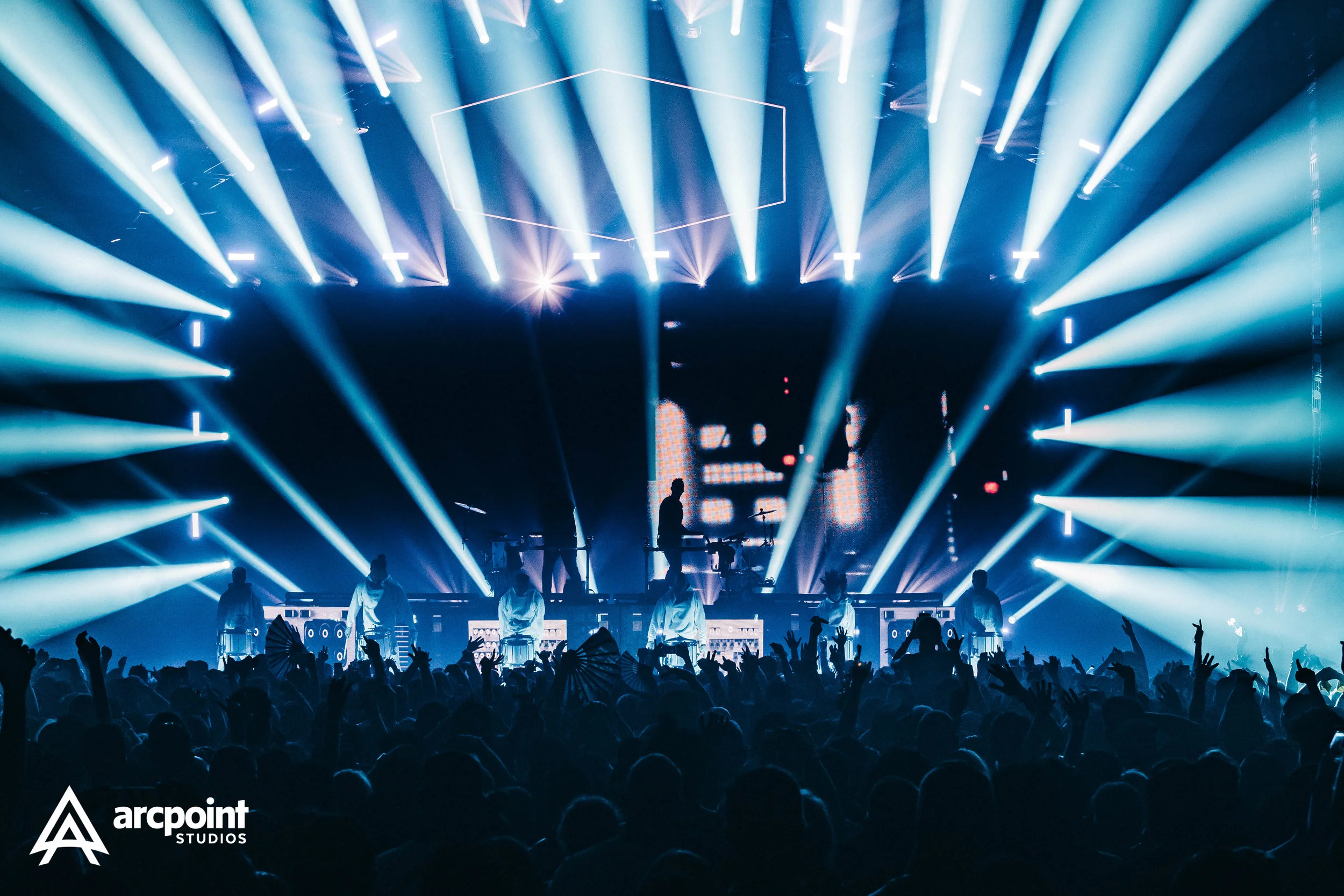 A concert stage illuminated with bright blue and white lights, showing silhouettes of performers and a DJ behind a setup, with an audience raising hands in front.