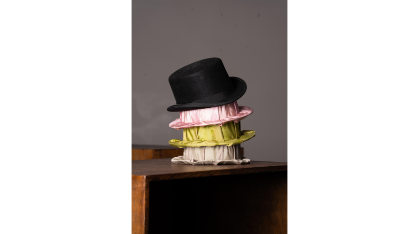 Stack of hats on a wooden surface: black top hat placed over layered pink, green, and beige fabric hats.
