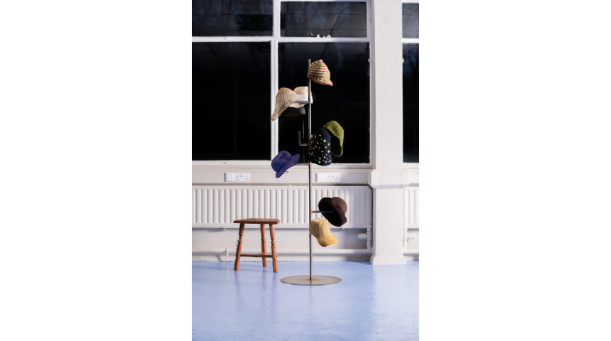 Freestanding metal hat stand displaying multiple colourful hats; small wooden stool beside it, set against large windows at night.