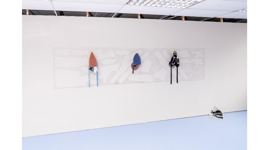 Three handmade hats mounted on a long wall drawing; pale sketched background with sculptural textile hats in orange, blue, and black.