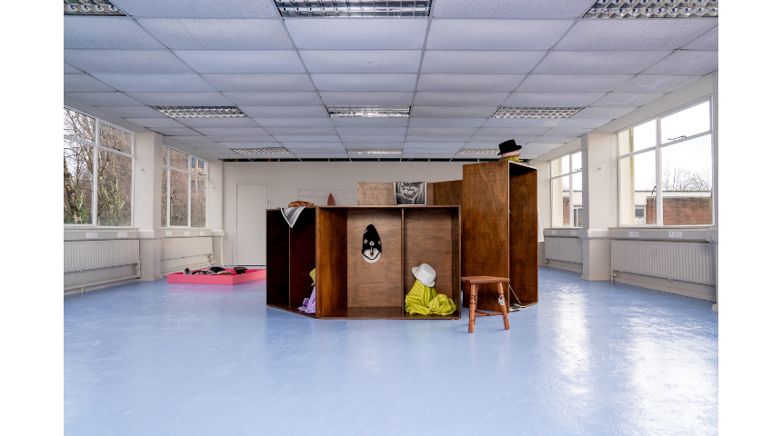Wide gallery view with blue floor and large windows; central wooden display structure holds hats, fabrics, and objects arranged in compartments.