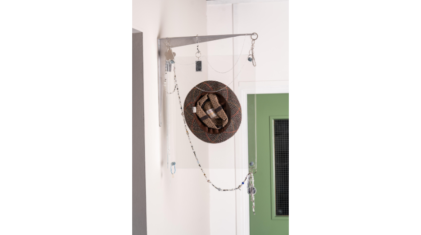 Single hat suspended on a metal bracket with chains and jewellery; checked fabric hat displayed against a white wall and green door.