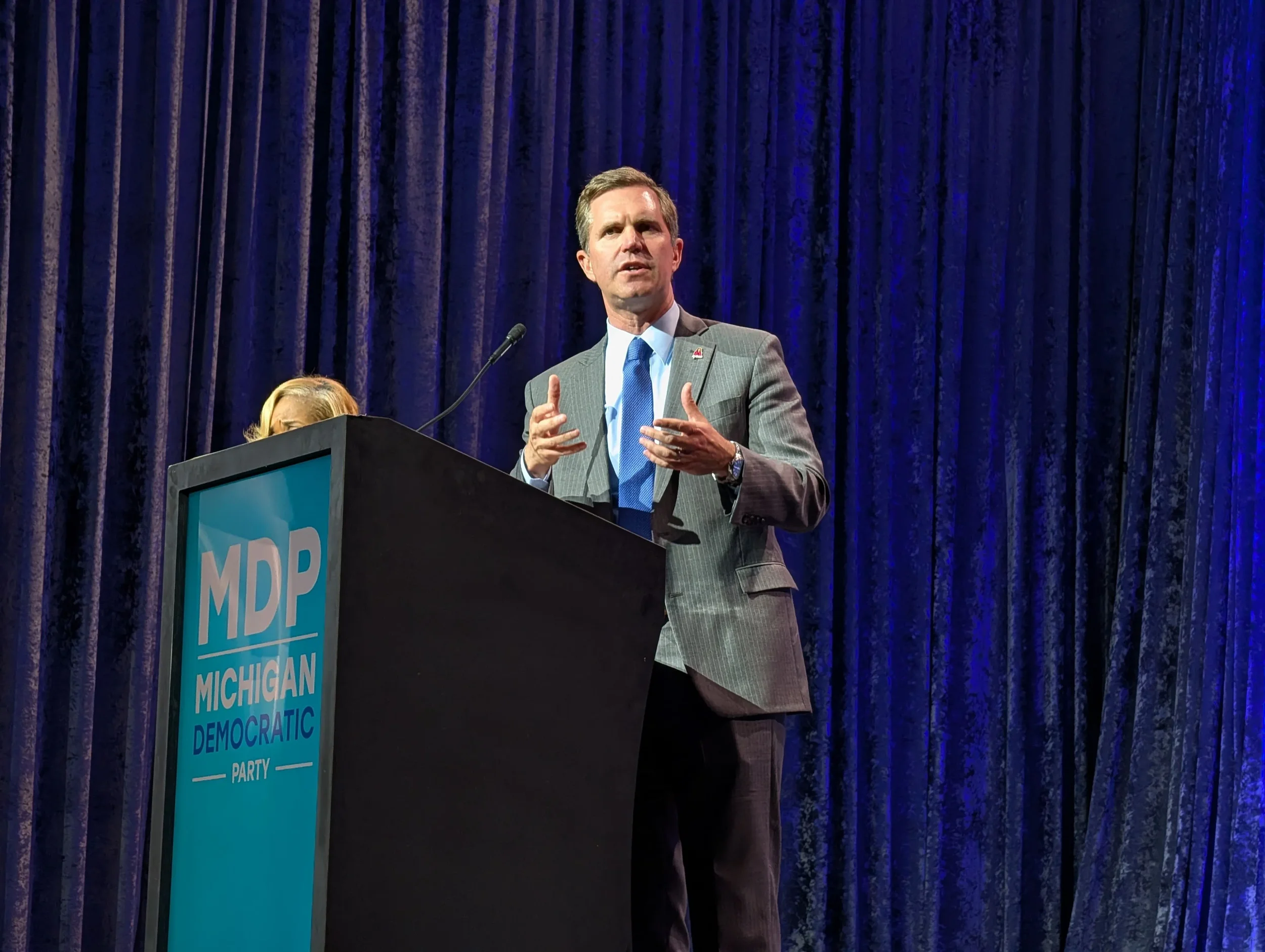 Andy Beshear delivering a speech in front of a blue backdrop at the Michigan Democratic Party convention