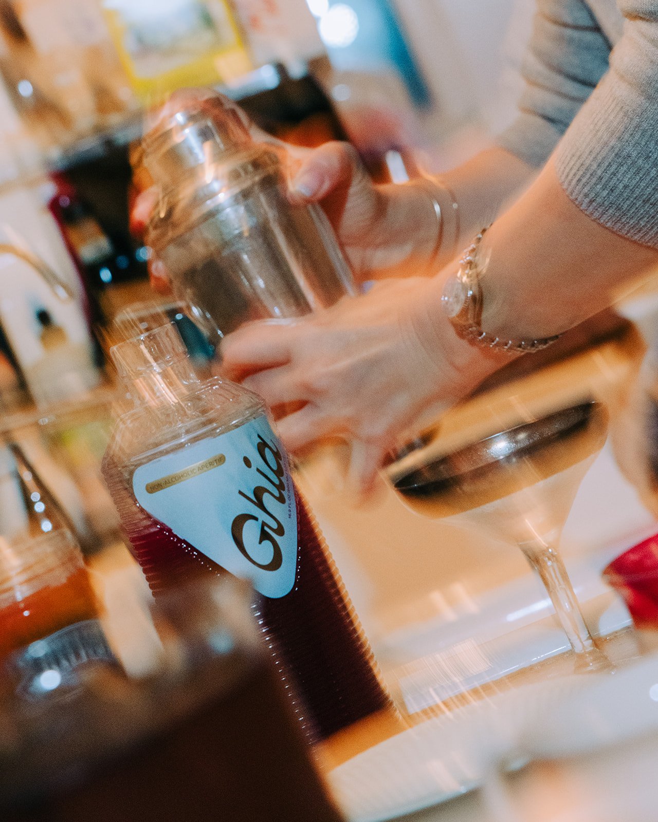 Person holding a cocktail shaker near a glass with a dark drink, inside a bar or kitchen setting with various bottles and jars in the background.
