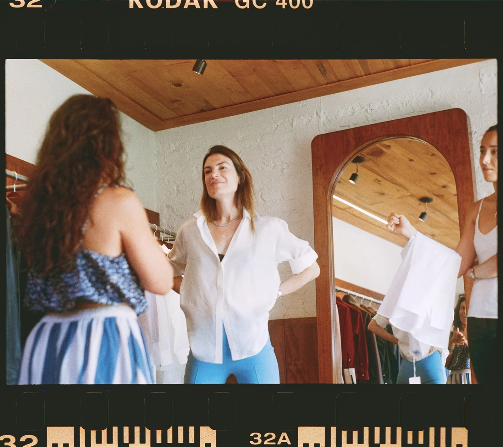 Three women in a clothing store, one trying on clothes in front of a mirror, the other two helping or observing, with racks of clothes in the background.