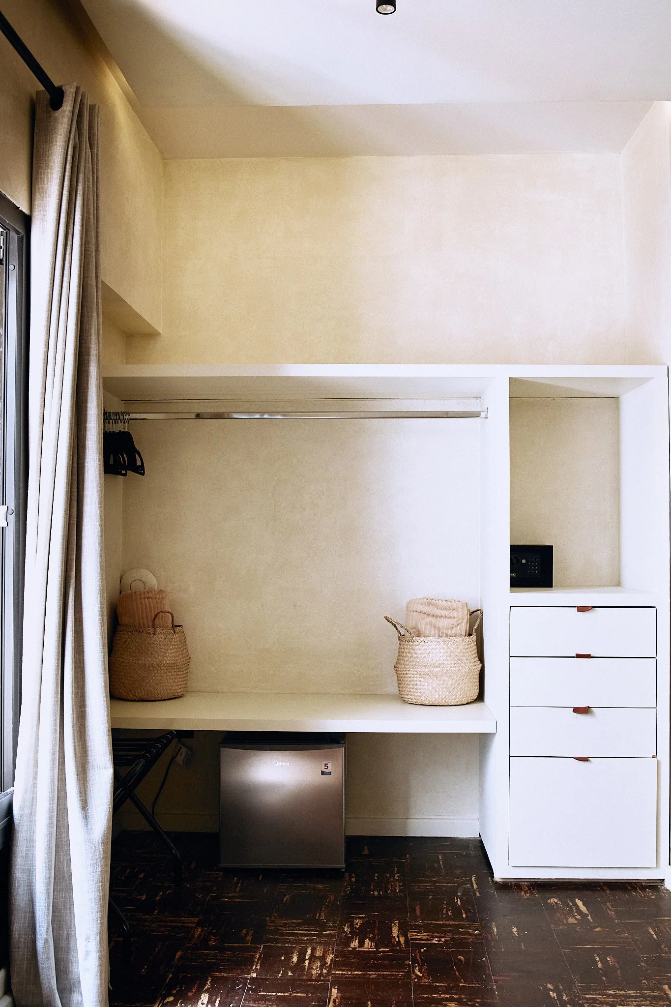 Empty walk-in closet with beige walls, a hanging rod, two woven baskets, a mini-fridge, and a set of white drawers.