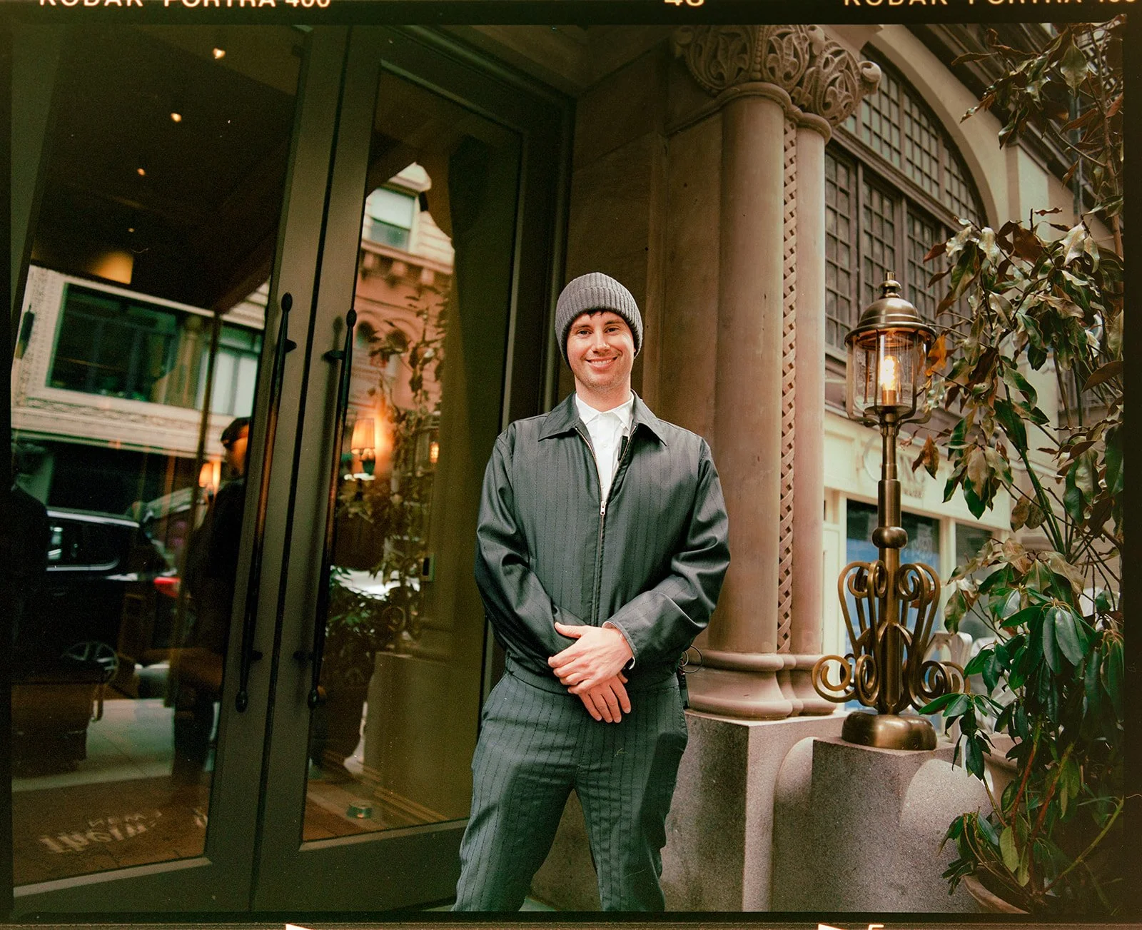 A man standing outdoors near a building entrance, smiling at the camera, wearing a gray beanie, a striped suit, and a dark jacket, with decorative architectural elements and plants in the background.