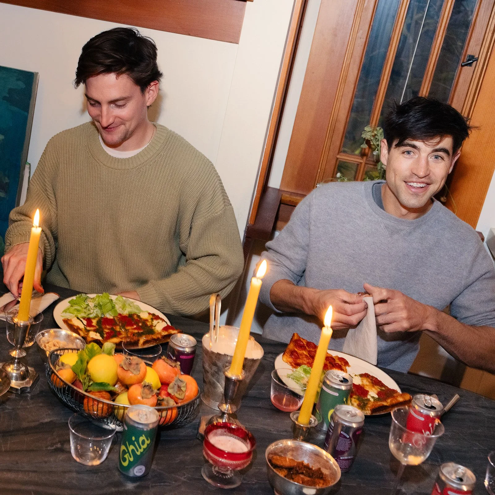 Two young men sit at a dinner table with lit candles, pizza, fruit, soda cans, and snacks, smiling and preparing to eat.