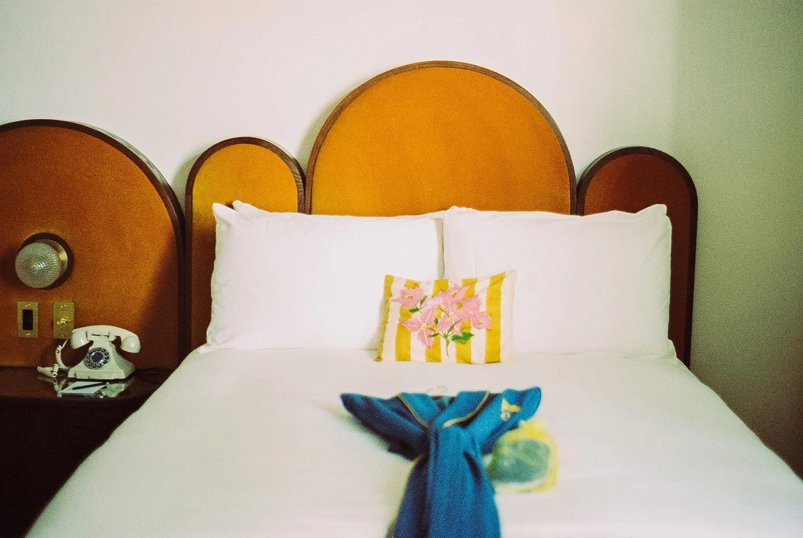 Hotel bed with white pillows, a decorative pillow with pink flowers and yellow stripes, and a blue towel. There is a wooden headboard with a rounded design, and a nightstand with a white landline phone and a wall-mounted light or speaker.