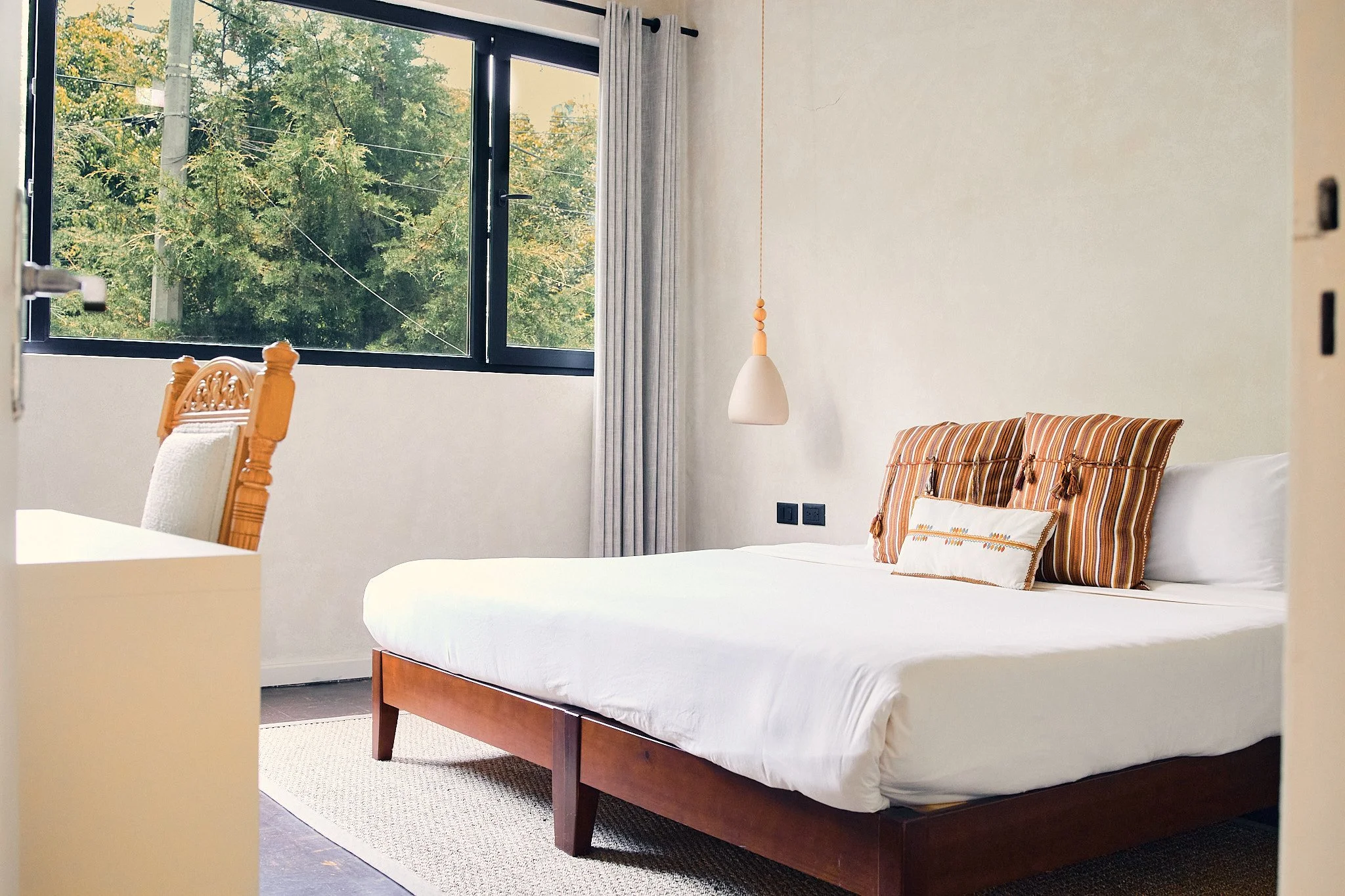 A bedroom with a bed, striped pillows, a large window with gray curtains, and a pendant light hanging from the ceiling.