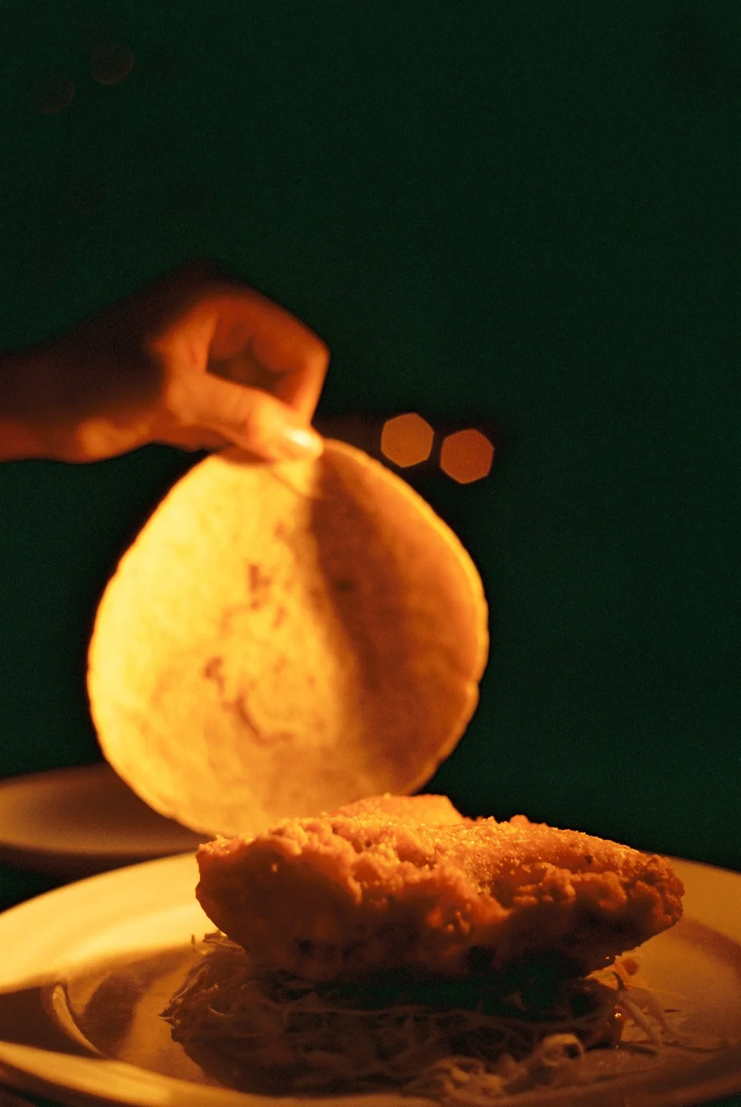 A hand holds a lit candle over a plate with a piece of bread, with a dark background and two blurred hexagonal light reflections.