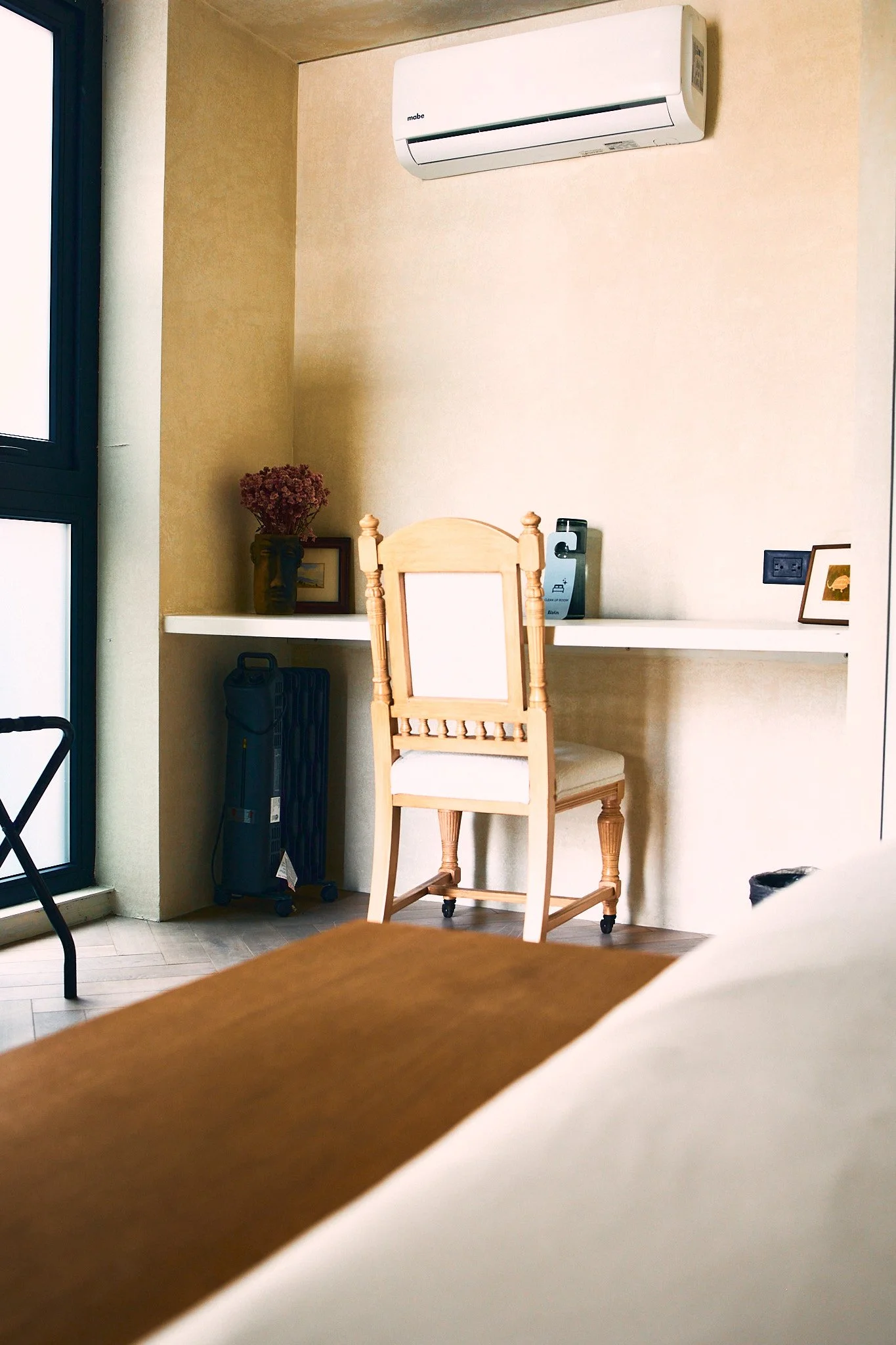 A corner of a room with a window, beige walls, a white air conditioning unit mounted on the wall, a small shelf with a vase of flowers, a framed picture, a utility box, a black power outlet, wooden furniture, and part of a wooden table in the foregro