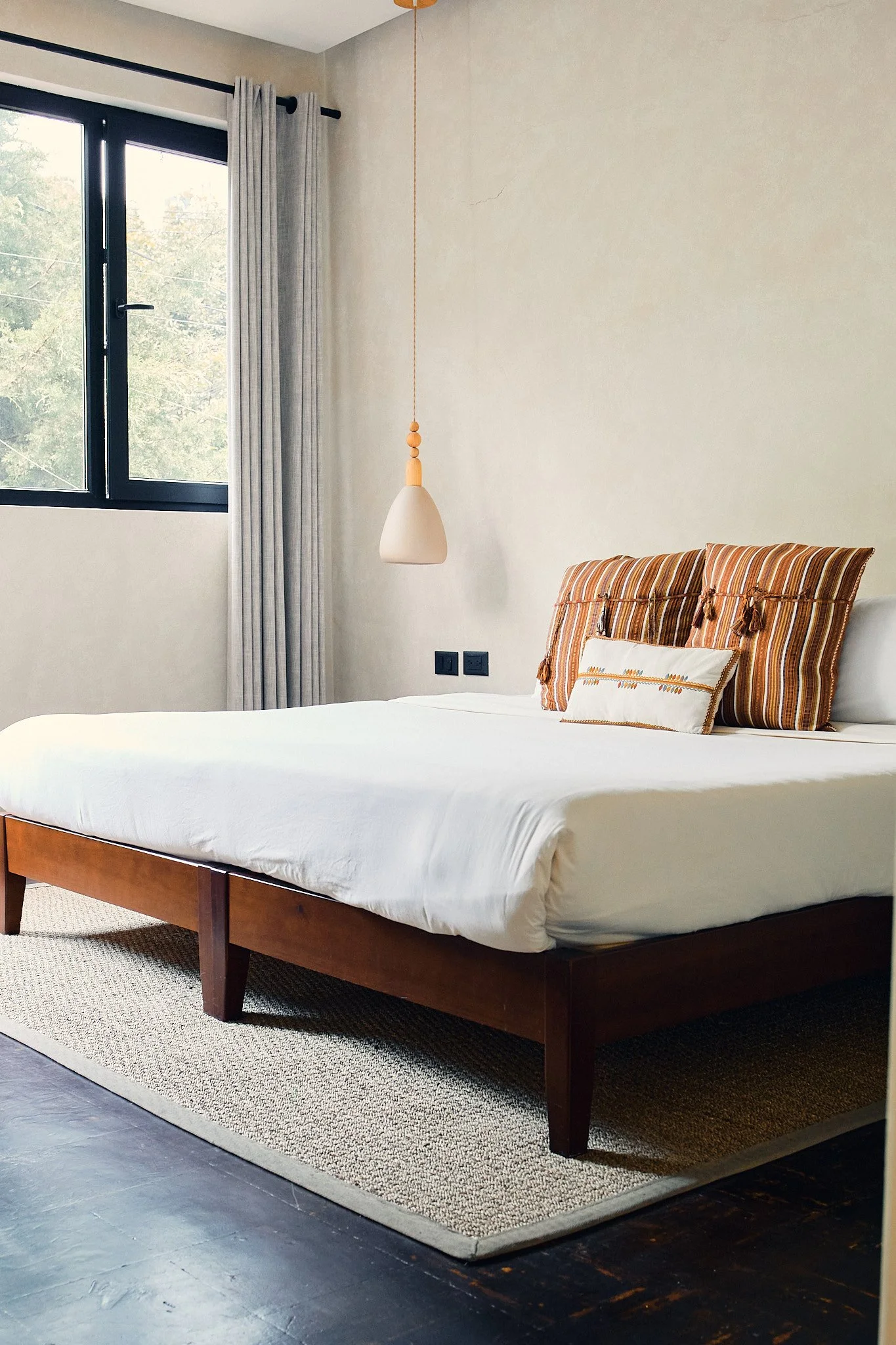 A bedroom with a bed featuring white bedding, two decorative pillows with striped patterns, and a white pillow with orange embroidery. There is a window with gray curtains, a hanging pendant light, and a beige rug on a dark wooden floor.