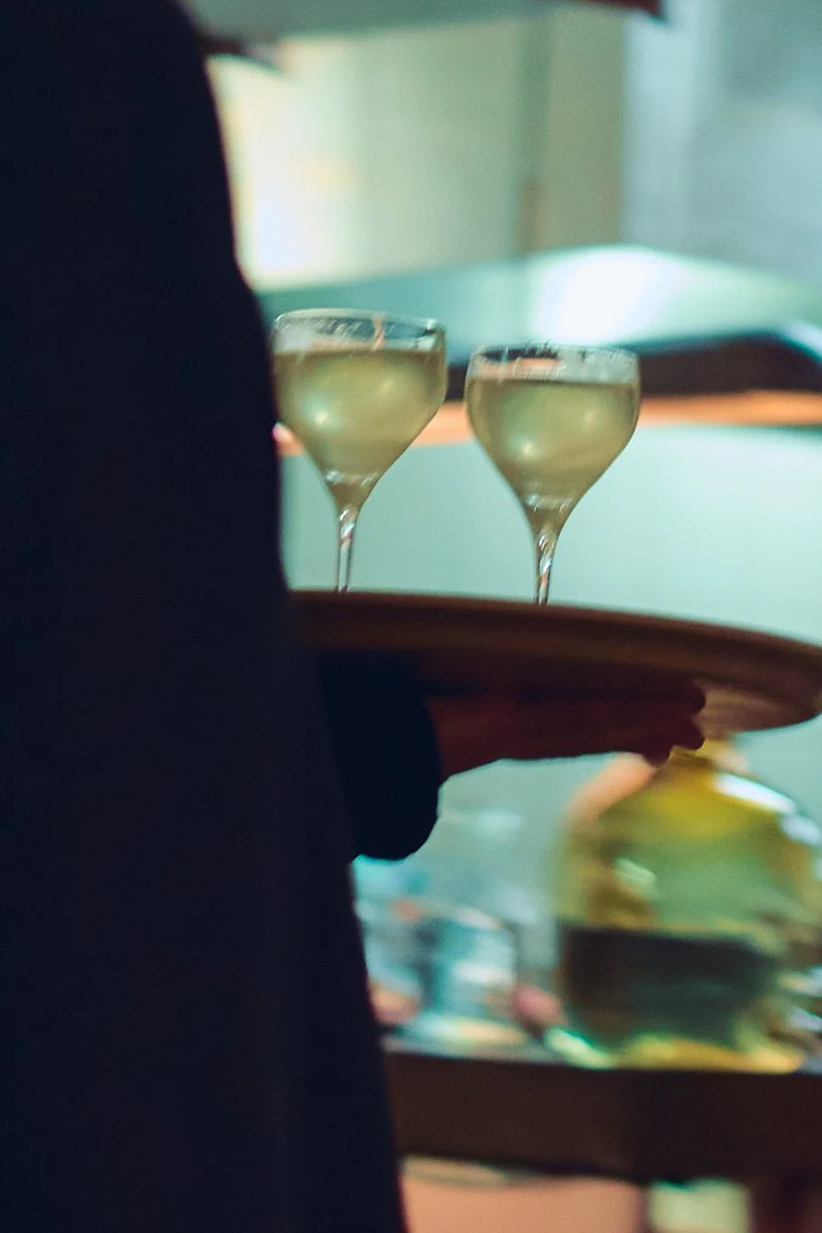 A person holding a wooden tray with two glasses of white wine in a dimly lit setting.