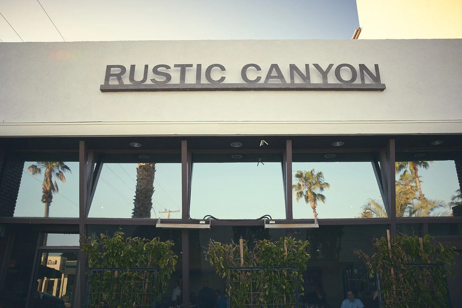 The storefront sign reads 'Rustic Canyon' with a view of palm trees and a clear sky in the background.
