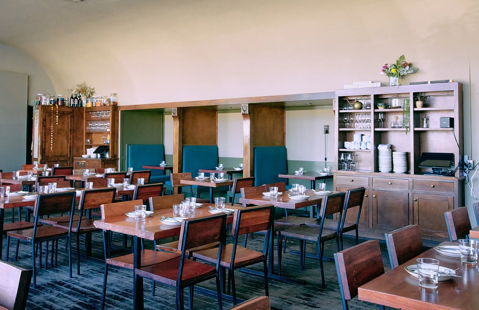 Empty restaurant dining room with wooden tables and chairs, a serving station with dishes and glasses, and a hutch with glassware and plates on the right.