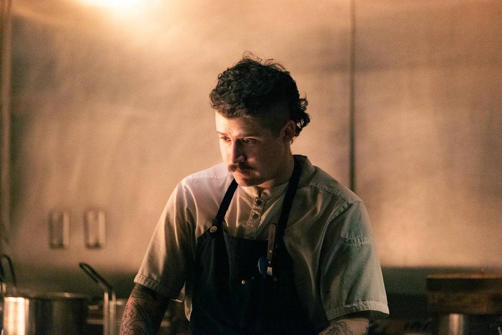A man with dark hair, a mustache, and tattoos on his arms is wearing a beige button-up shirt and a black apron. He appears to be working in a kitchen with a focused or contemplative expression, dim lighting creating a warm ambiance.