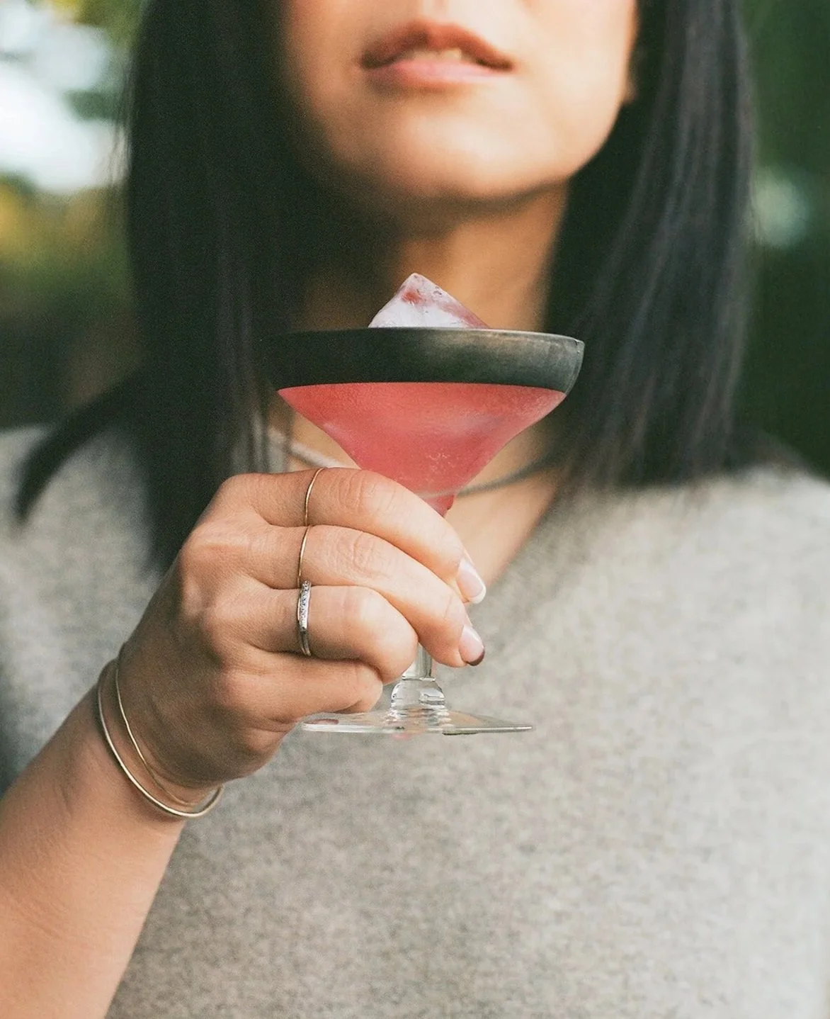 A woman holding a pink cocktail glass with a slice of ice, wearing rings and bracelets