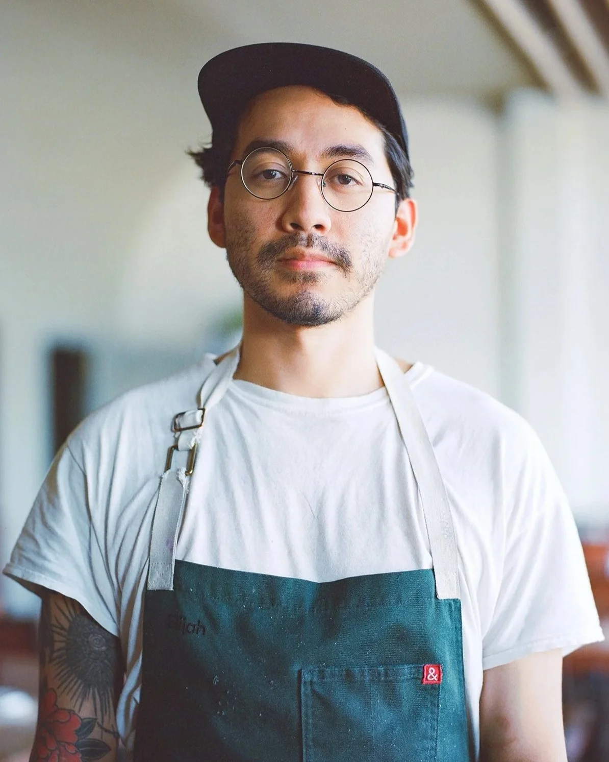 A man with glasses, a black cap, a white T-shirt, and a black apron with a pocket, standing indoors with a neutral background.