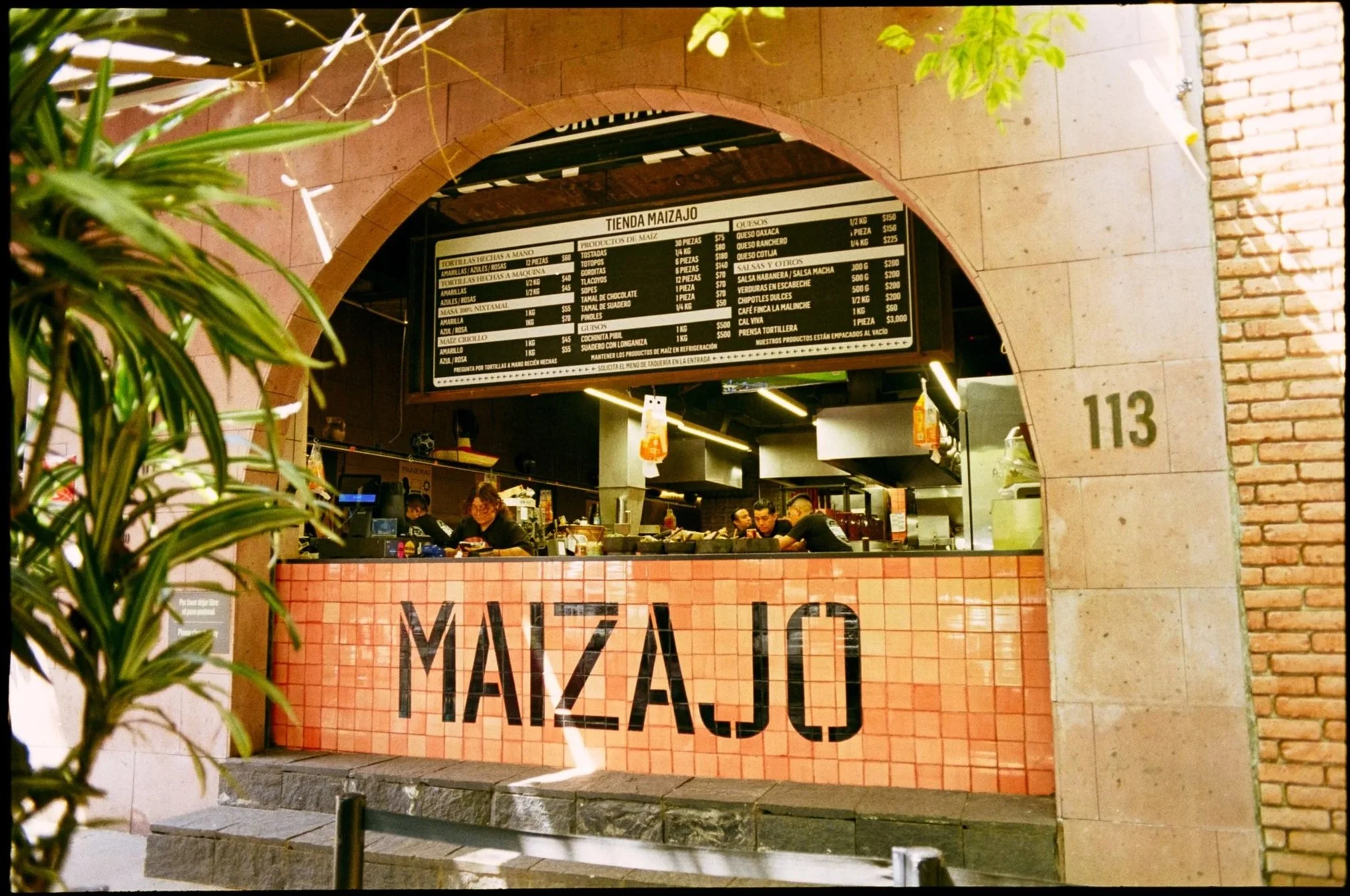 Close-up of a digital display or menu board at a food establishment, with various menu items and prices listed in Spanish, and part of the entrance or facade with brick and stone details visible.