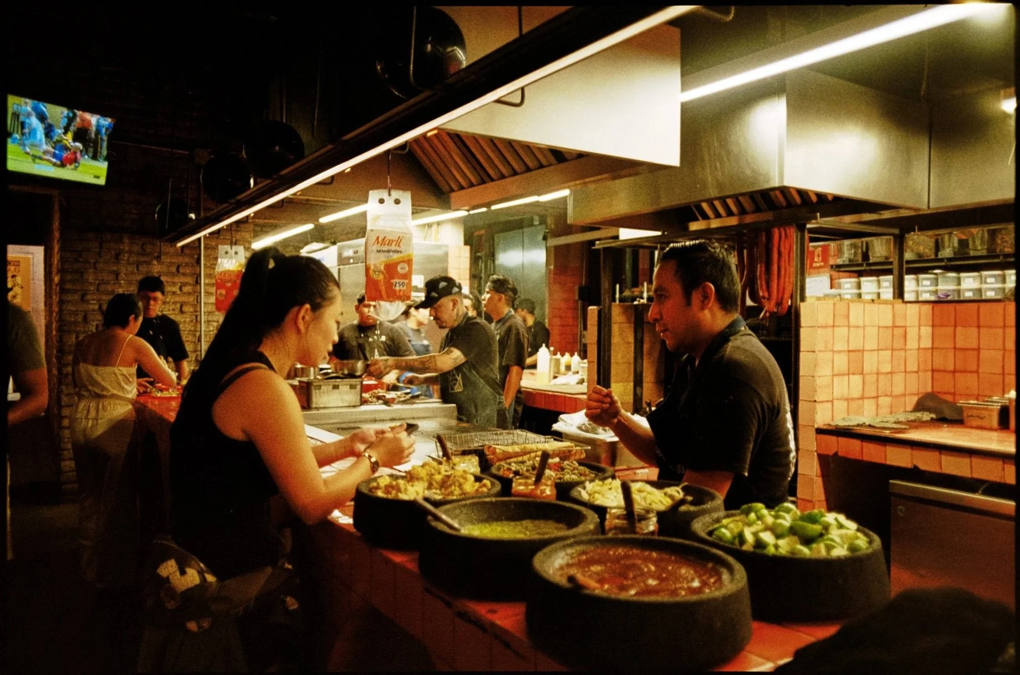 A bustling restaurant or food market with people ordering and serving food. Several bowls of food, including vegetables and sauces, are visible on the counter. The background features a brick wall, hanging signs, and a television displaying a sports 
