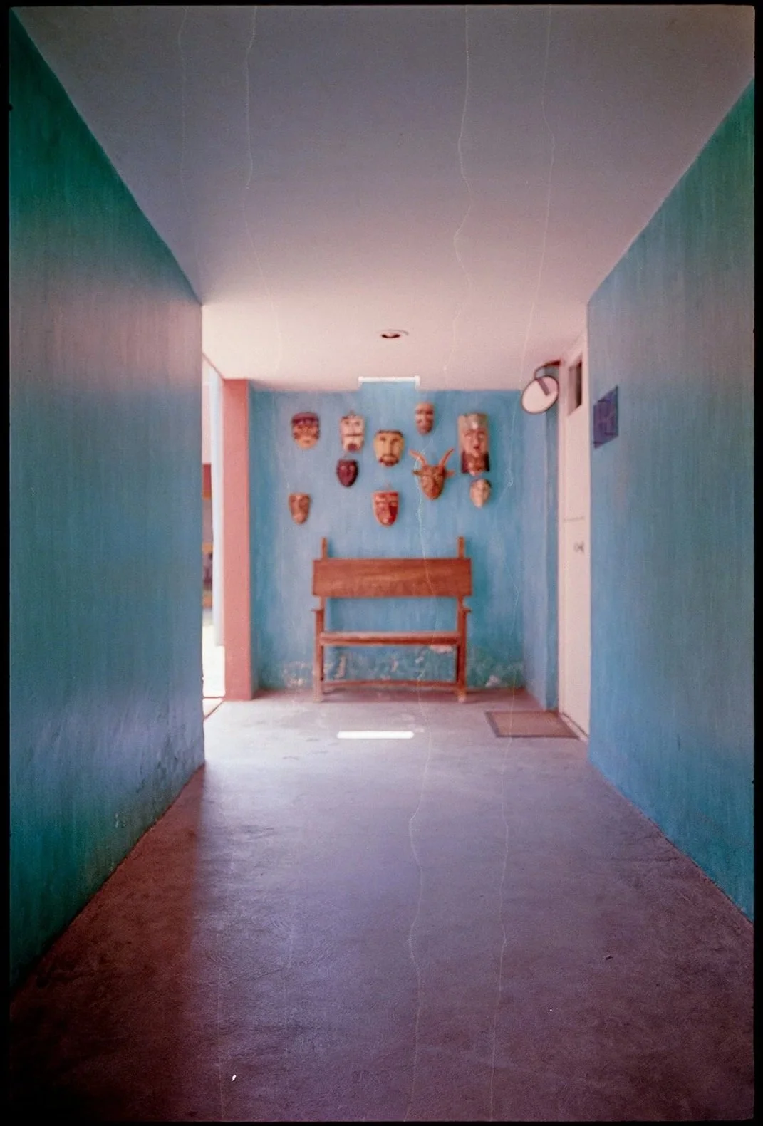 A hallway with blue painted walls leads to a wall decorated with various masks and a mounted deer head, a wooden bench below the decoration, and a door on the right side.