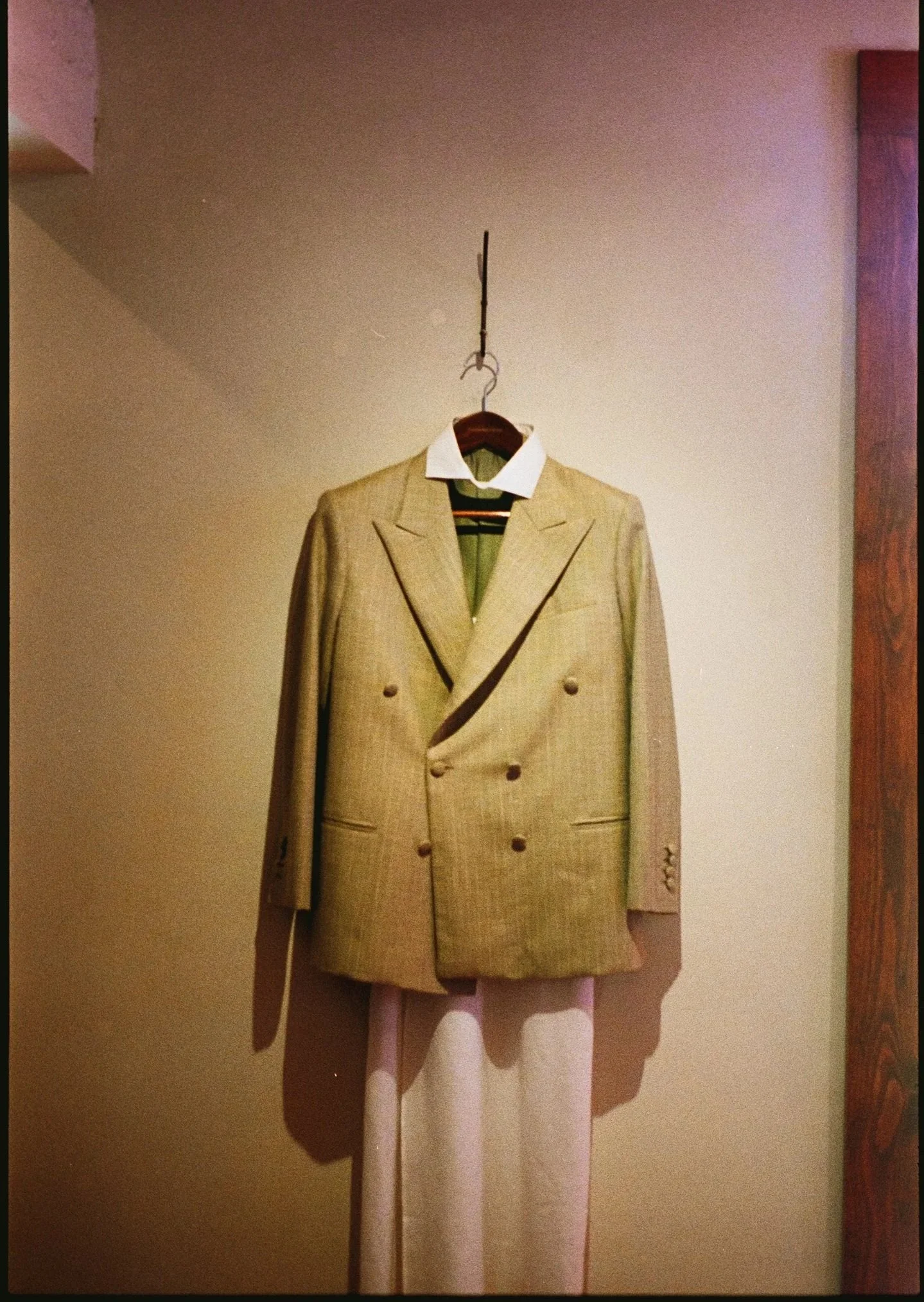 A vintage men's suit jacket hanging on a hanger, with a collared shirt and sweater underneath. The jacket is beige with a checkered pattern and has double-breasted buttons. The outfit is displayed against a plain wall, with the person's legs visible 