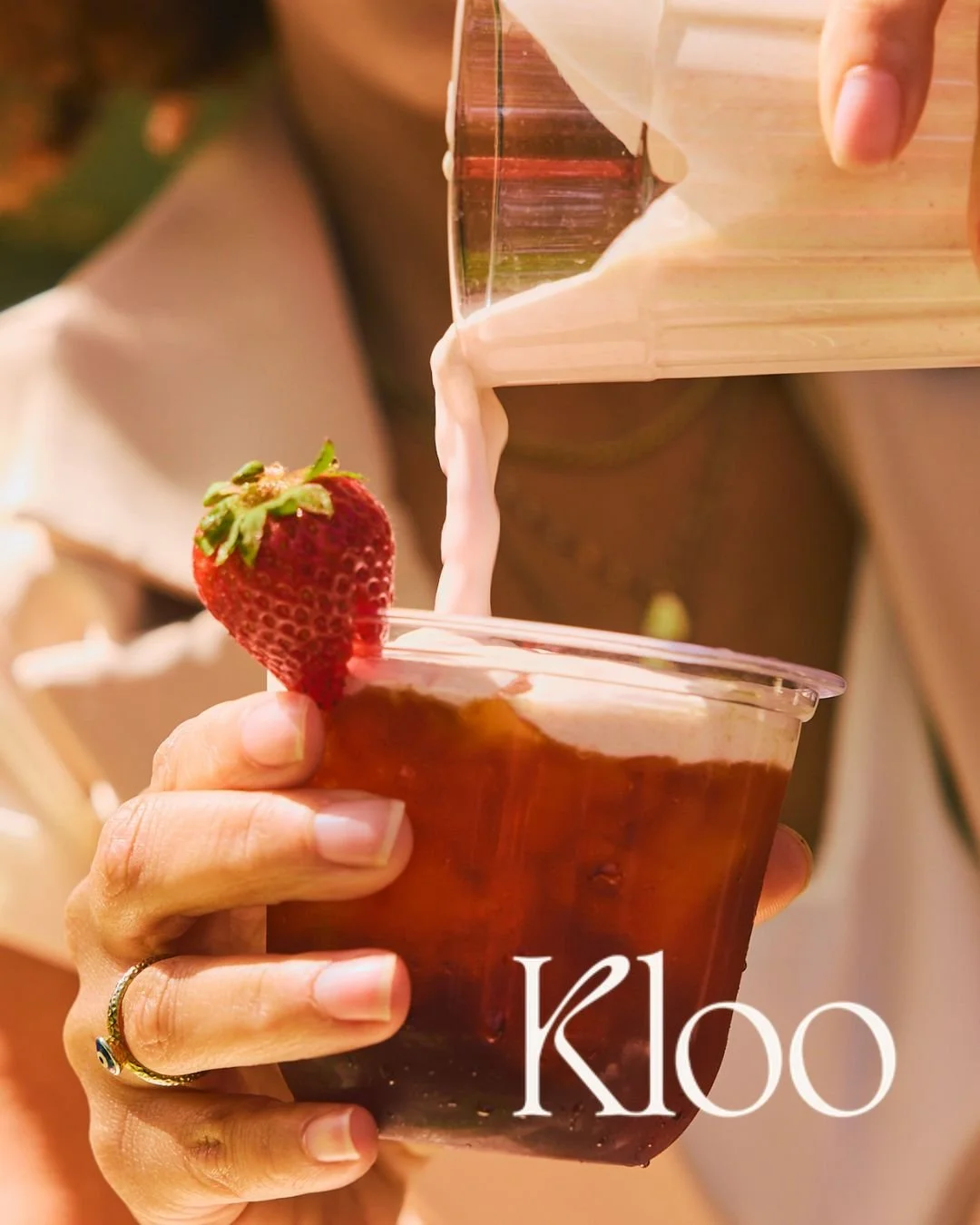 A person holding a plastic cup filled with iced tea, garnished with a strawberry on the rim, as cream is poured into the drink.