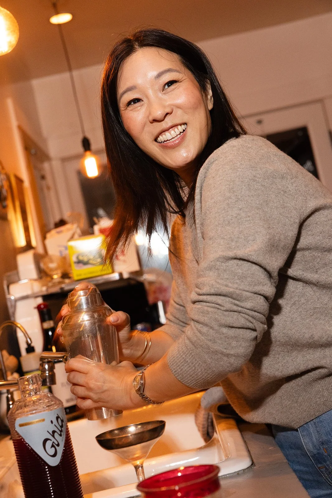A woman with black hair and a beige sweater smiling while preparing a cocktail in a kitchen.