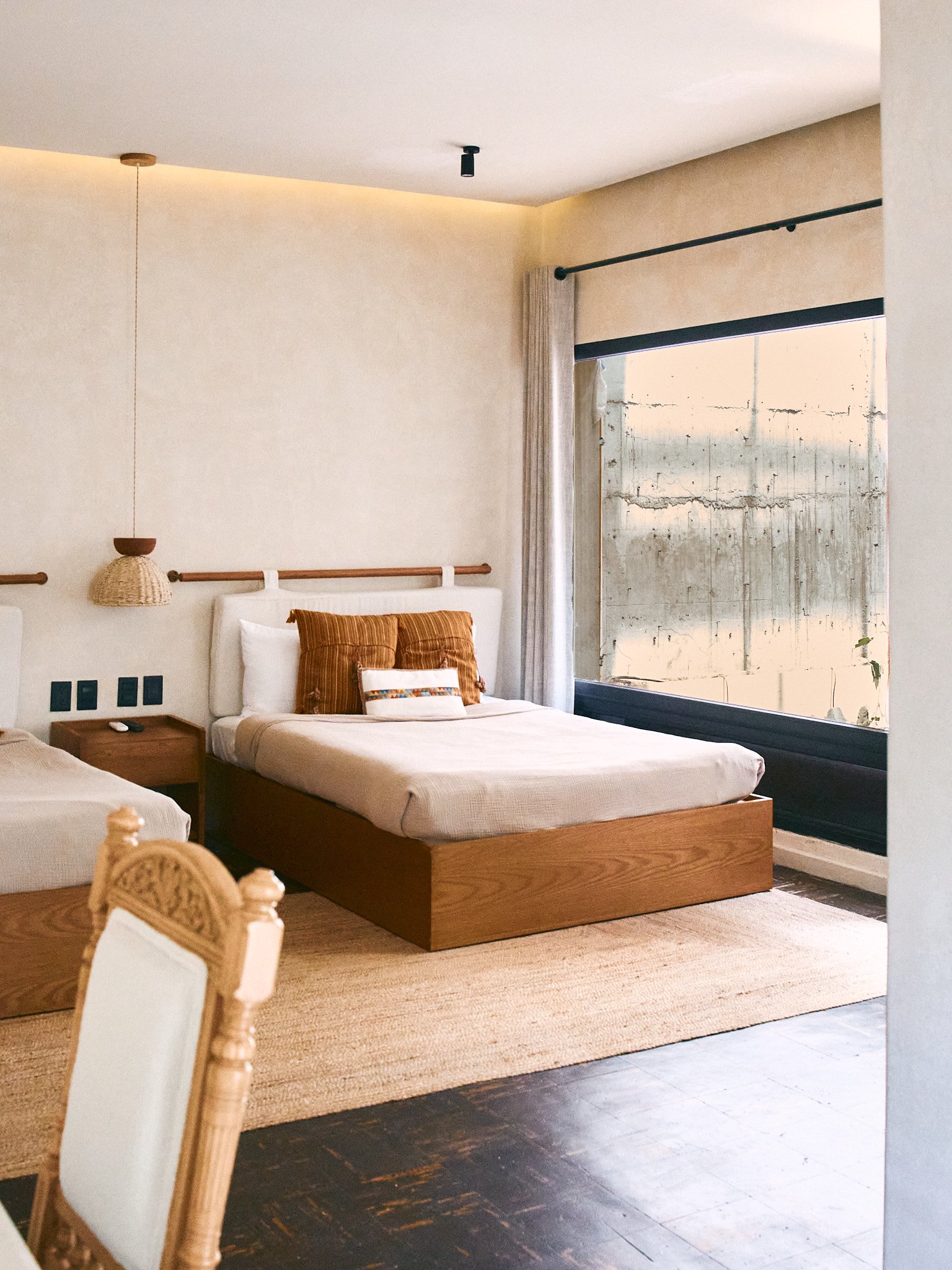 A bedroom with a bed featuring a white headboard and two rust-colored pillows, a wooden nightstand with a remote control, a hanging wicker lampshade, a large window with a black frame and a curtain, and a cement wall outside the window with exposed c