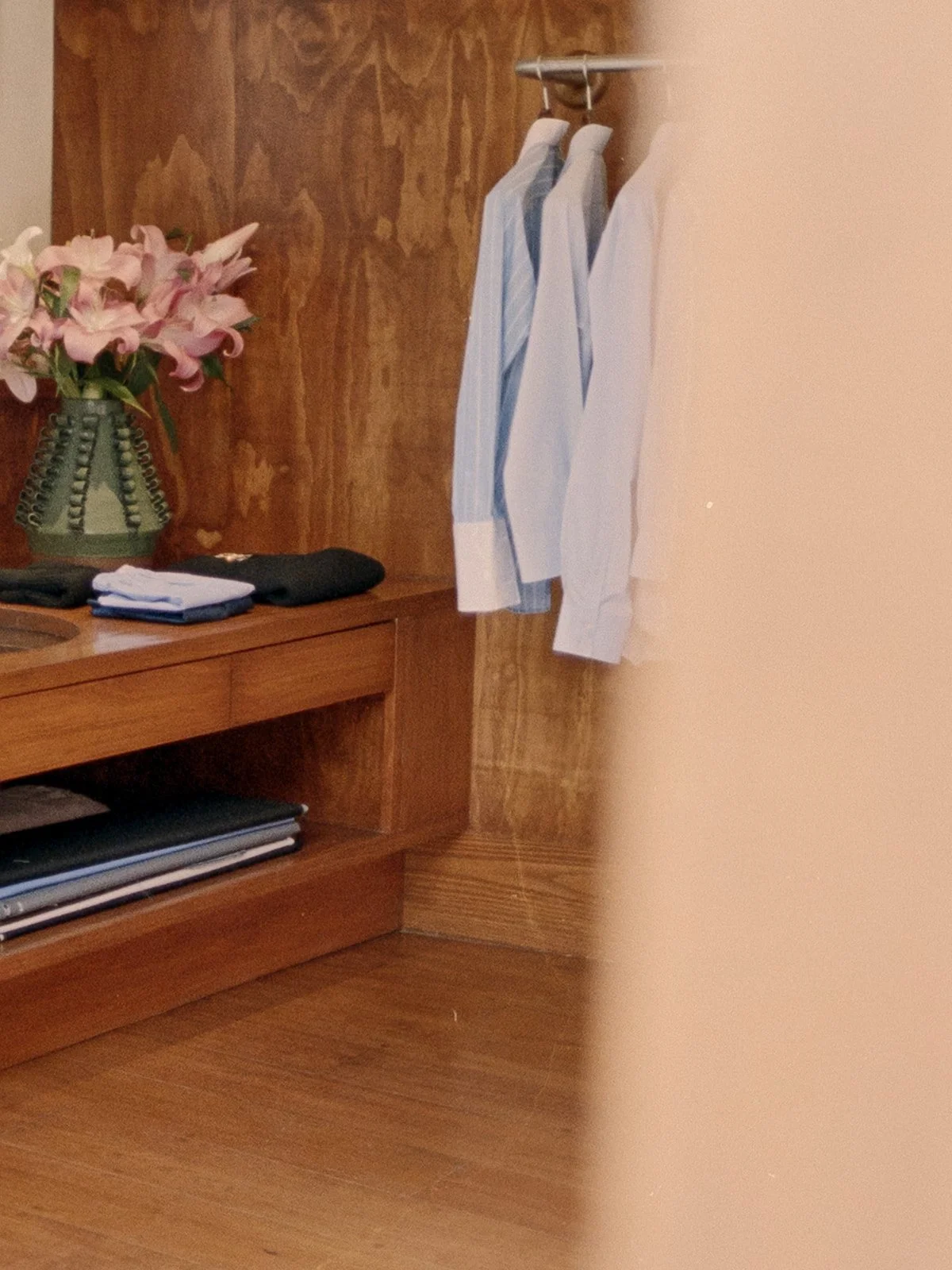 A wooden desk with a vase of pink flowers and neatly folded clothes, with hanging shirts on a rack beside a beige wall.