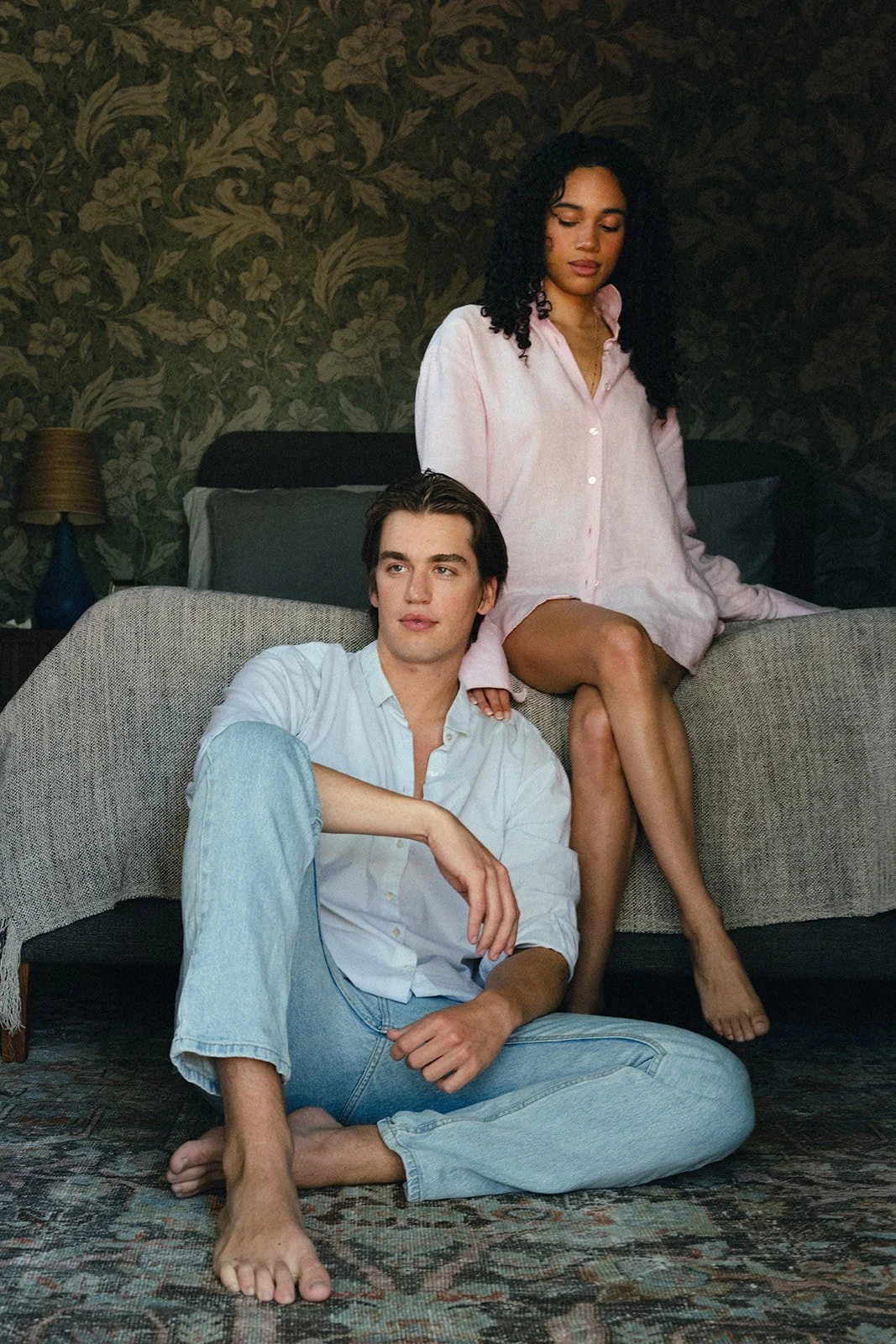 A man sitting on the floor with crossed legs, wearing a white shirt and light blue jeans, with a woman seated on a couch behind him, wearing a pink shirt dress, in a room with floral wallpaper.