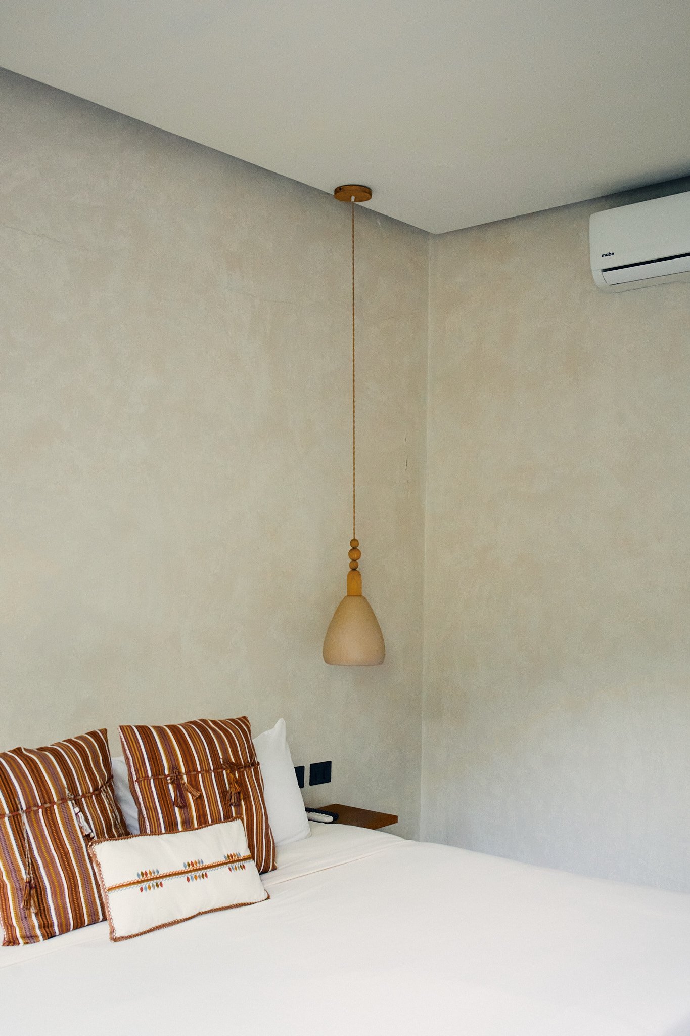 A bedroom with a white bed, striped pillows, wall-mounted beige lamp, air conditioning unit, and neutral-colored walls.