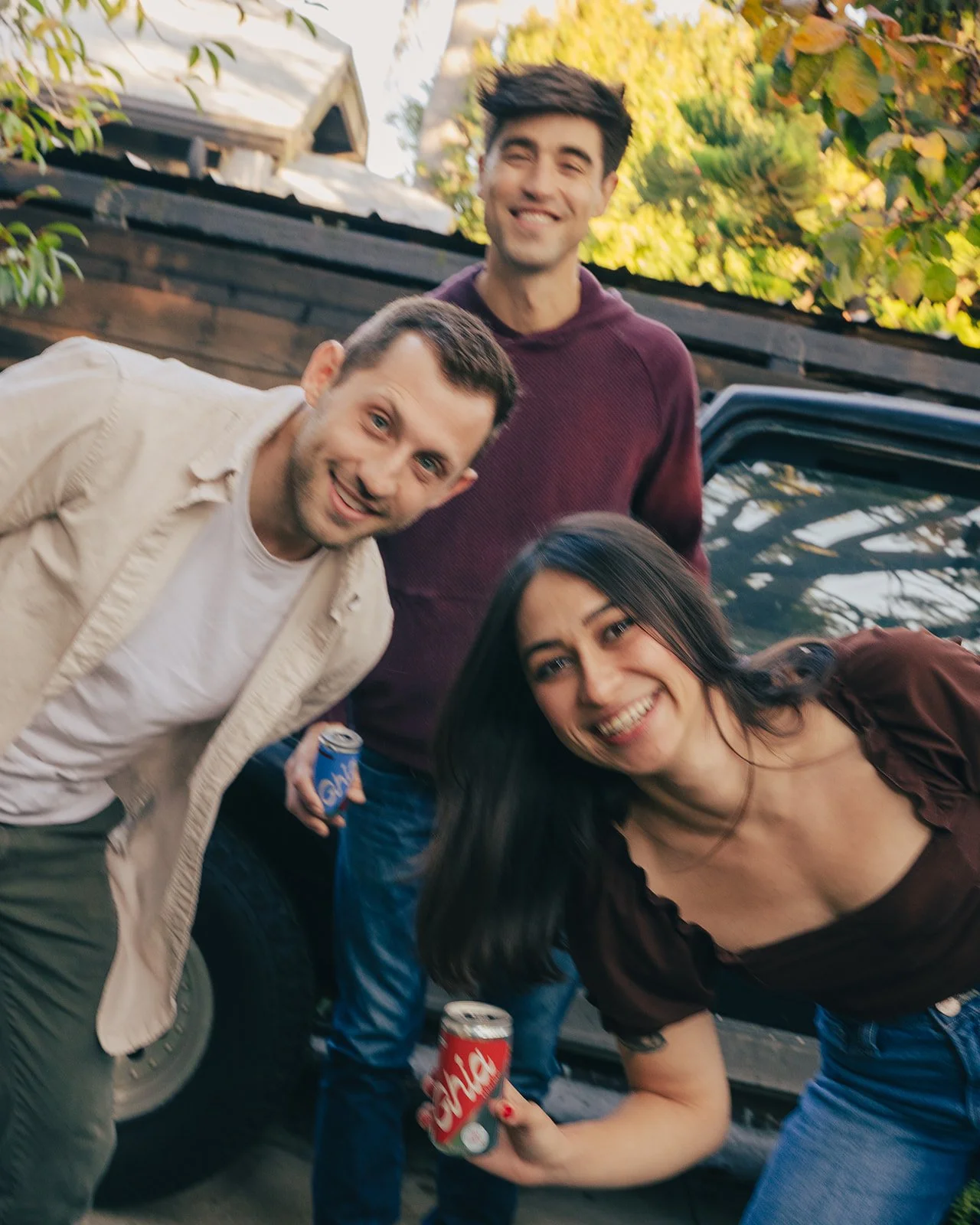 A group of four friends smiling and posing outdoors beside a black pickup truck. They are holding soda cans, with one person holding a can of Sprite and another a can of Coca-Cola. There are trees and a wooden fence in the background, indicating a casual outdoor gathering.