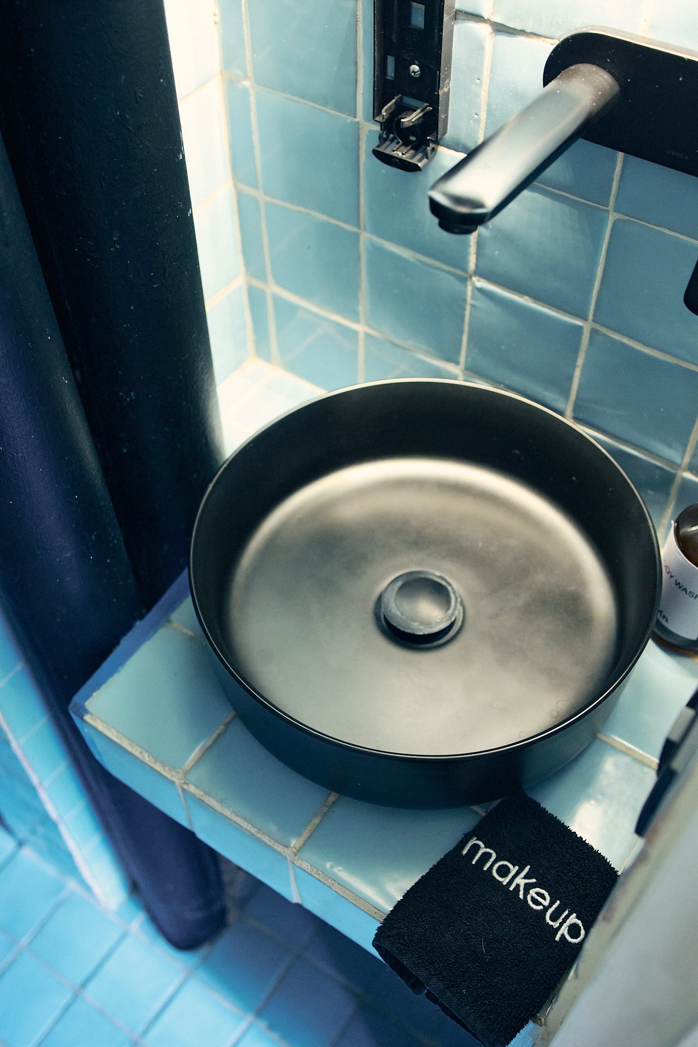 Close-up of a laundry machine with a metallic drum, positioned next to a black towel with the word 'makeup' embroidered on it, and a blue-tiled wall in the background.