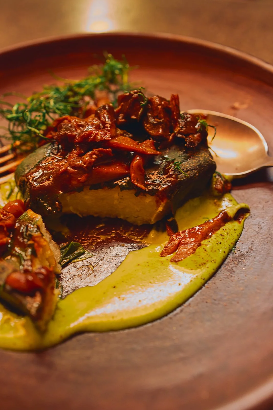 A plated dish featuring a grilled fish fillet topped with a beef and onion stew, garnished with herbs, served over a creamy green sauce.