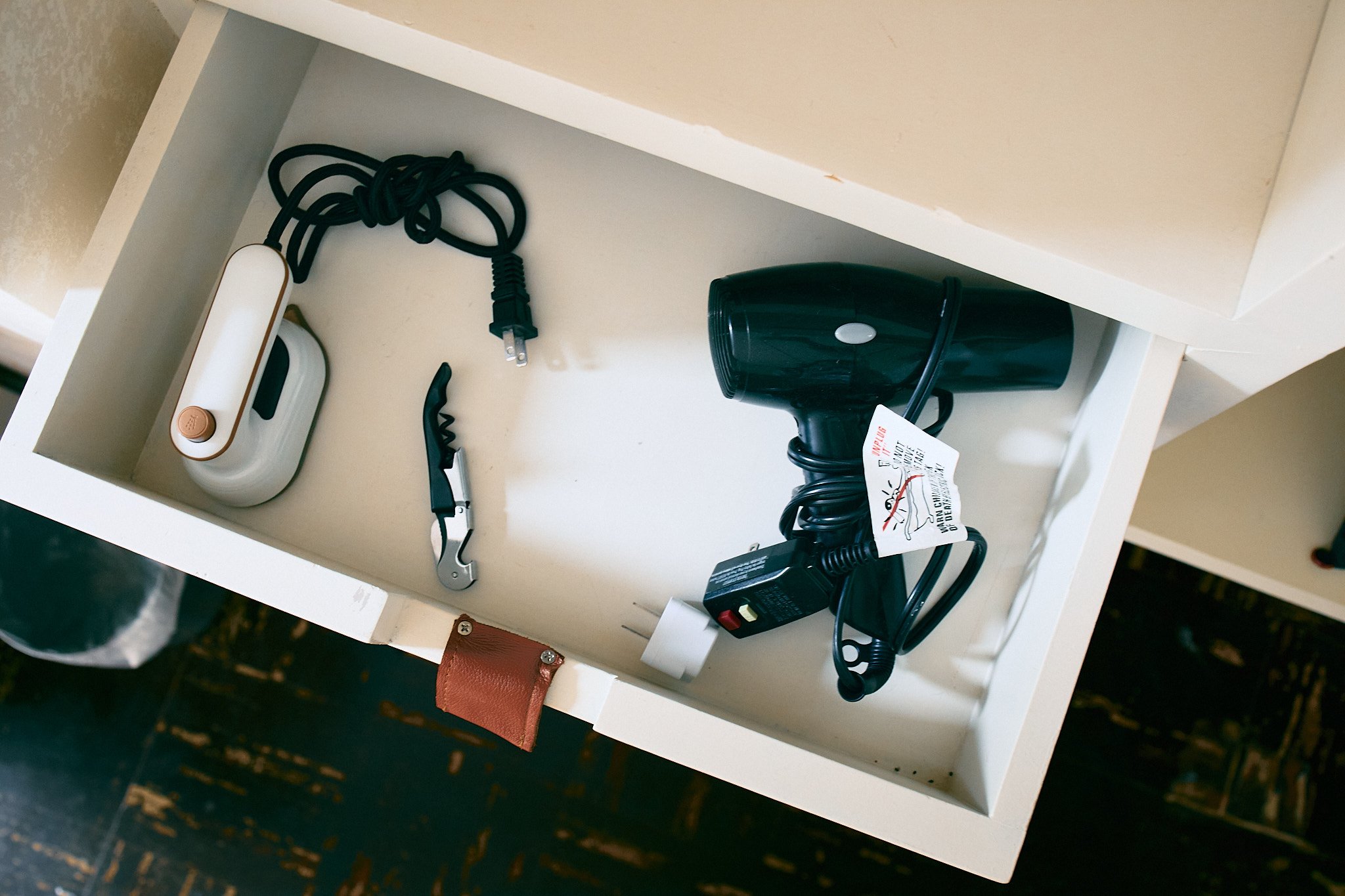The image shows a white shelf with a few electronic items and accessories, including a black hairdryer, a white and gold portable device, a pair of hair cutting scissors, a plugged-in power adapter, and an orange wallet or small bag.