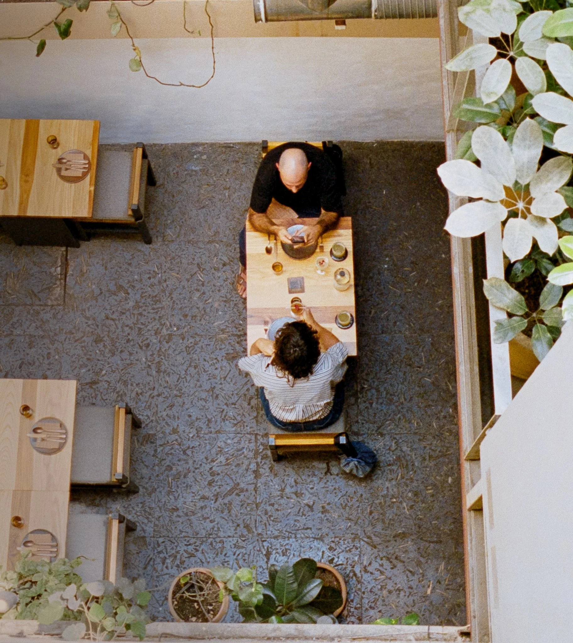 A man sitting at a small wooden table in a cafe, viewed from above, holding a smartphone. The table has various drinks and items on it. The setting appears cozy with plants around.