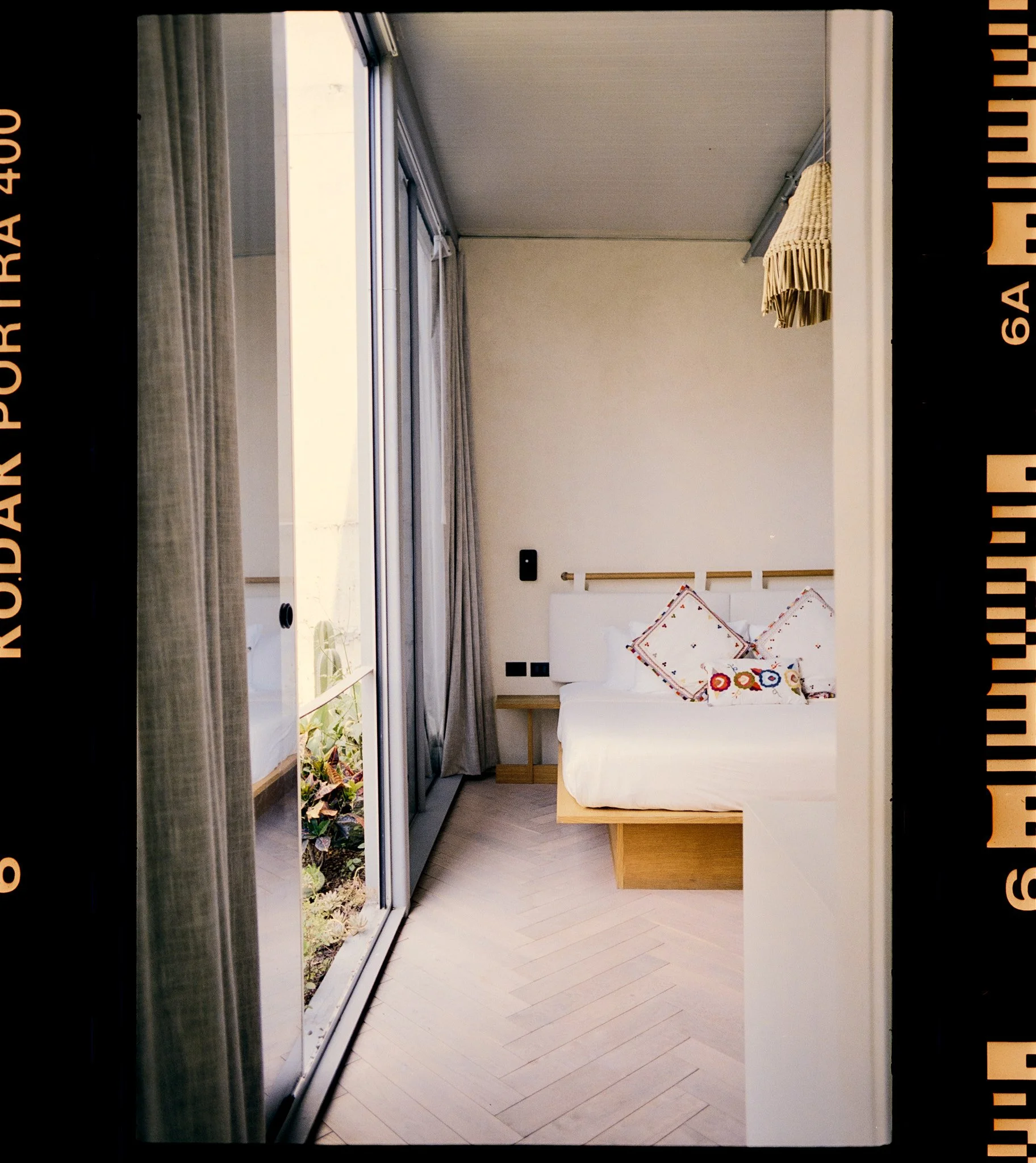 Interior view of a modern living space with a large sliding glass door leading to a balcony or outdoor garden area. The room has light-colored wooden flooring and a white couch with colorful patterned cushions. There is a small side table next to the