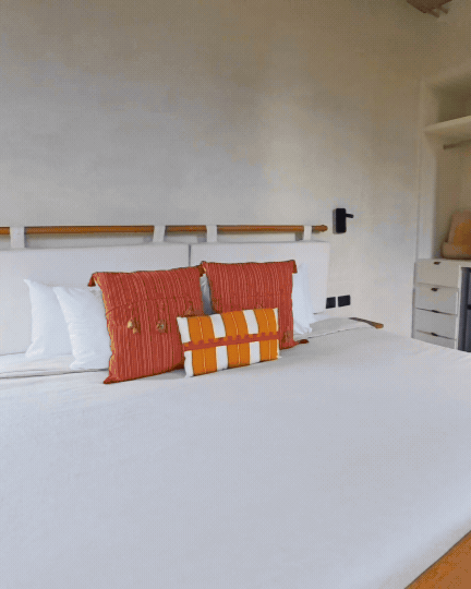 A neatly made bed with white sheets and orange striped pillows, in a minimalistic hotel room with a white wall and small bedside table.