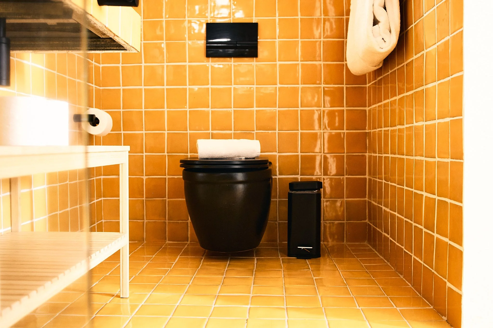 A small bathroom with orange tiled walls and floor, featuring a black toilet, a black trash can with a roll of toilet paper on top, a black tissue box holder, a white towel rolled up on a shelf, and a black flush button on the wall.